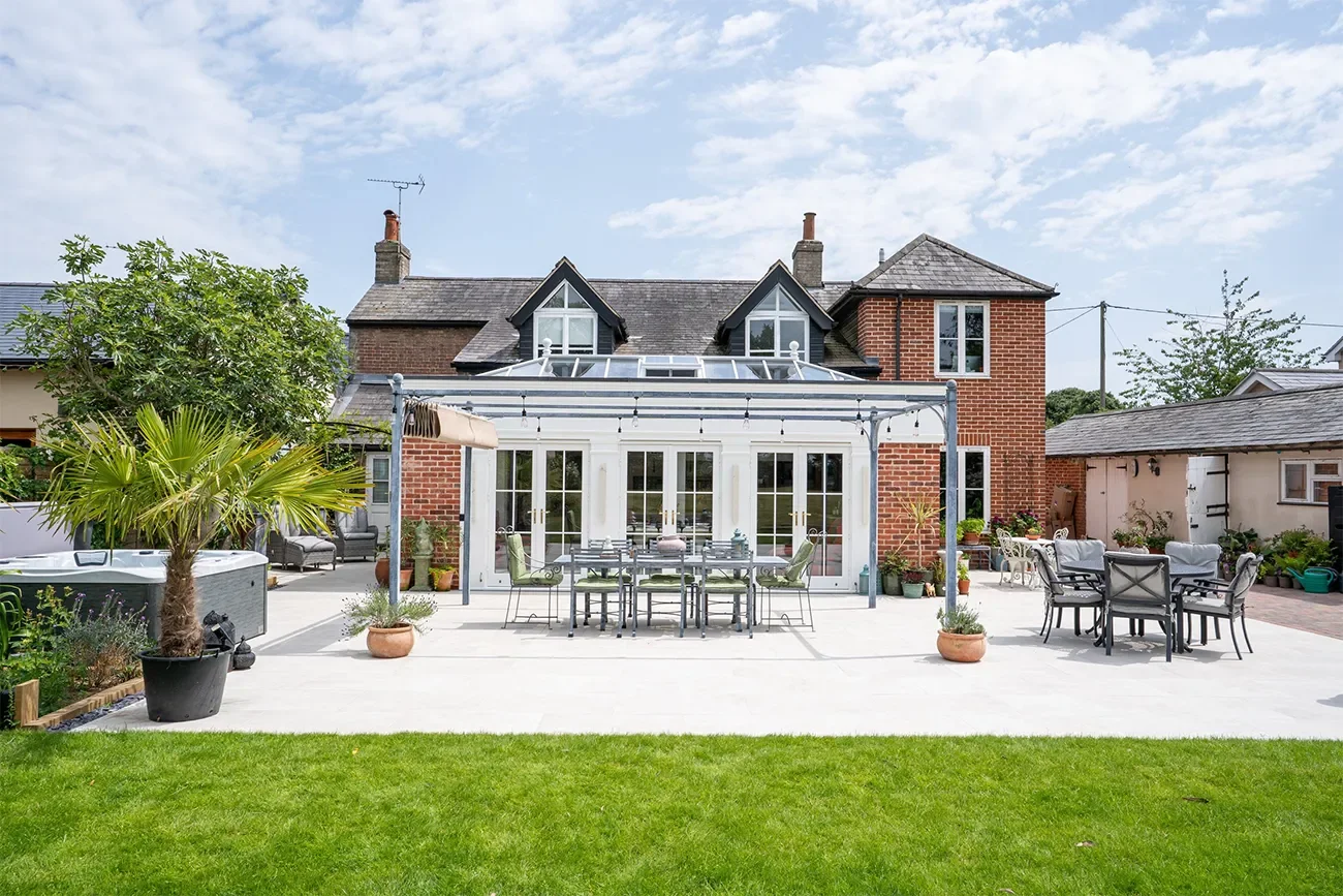 Backyard patio with outdoor dining table and chairs, potted plants, a hot tub, and a house with a brick facade and a glass conservatory