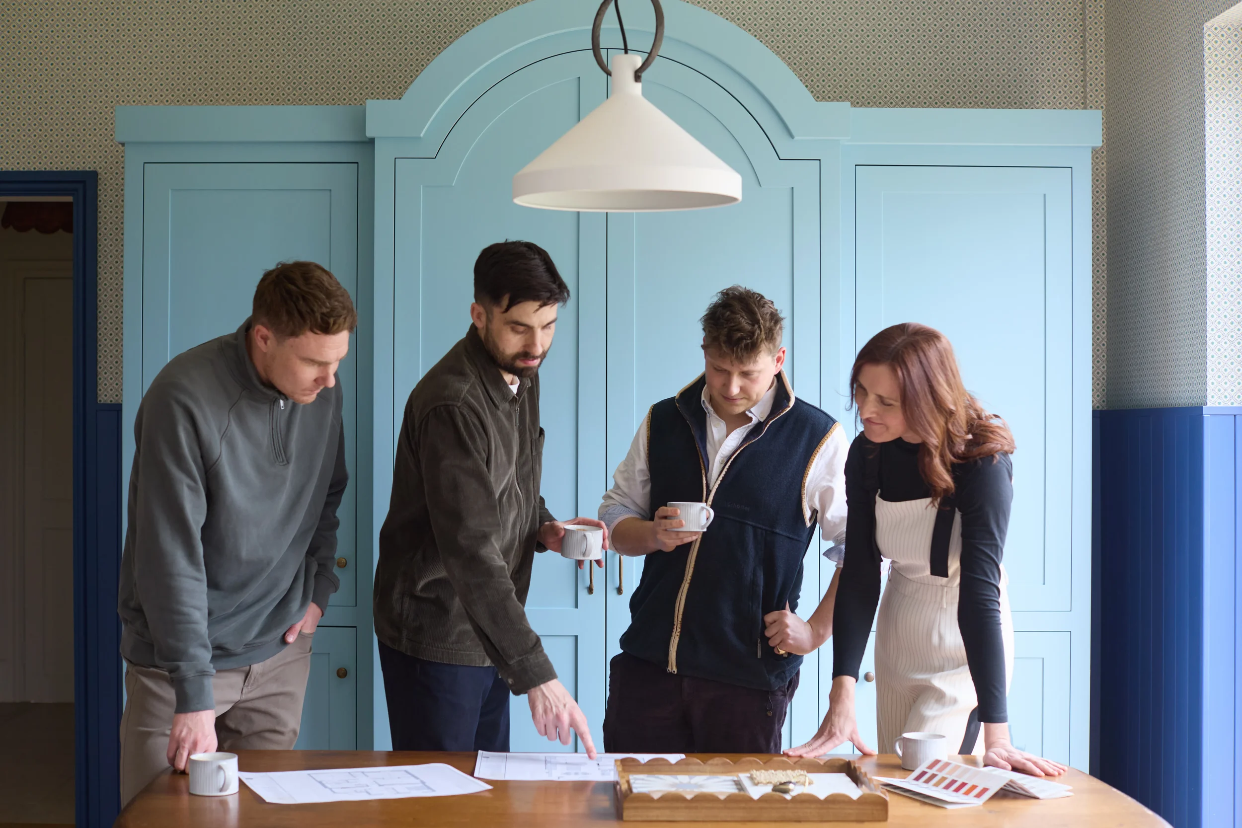 Four people gathered around a table looking at architectural plans or documents, in a room with a blue cabinet and patterned wallpaper. They are holding coffee mugs, and one person is pointing at the plans.