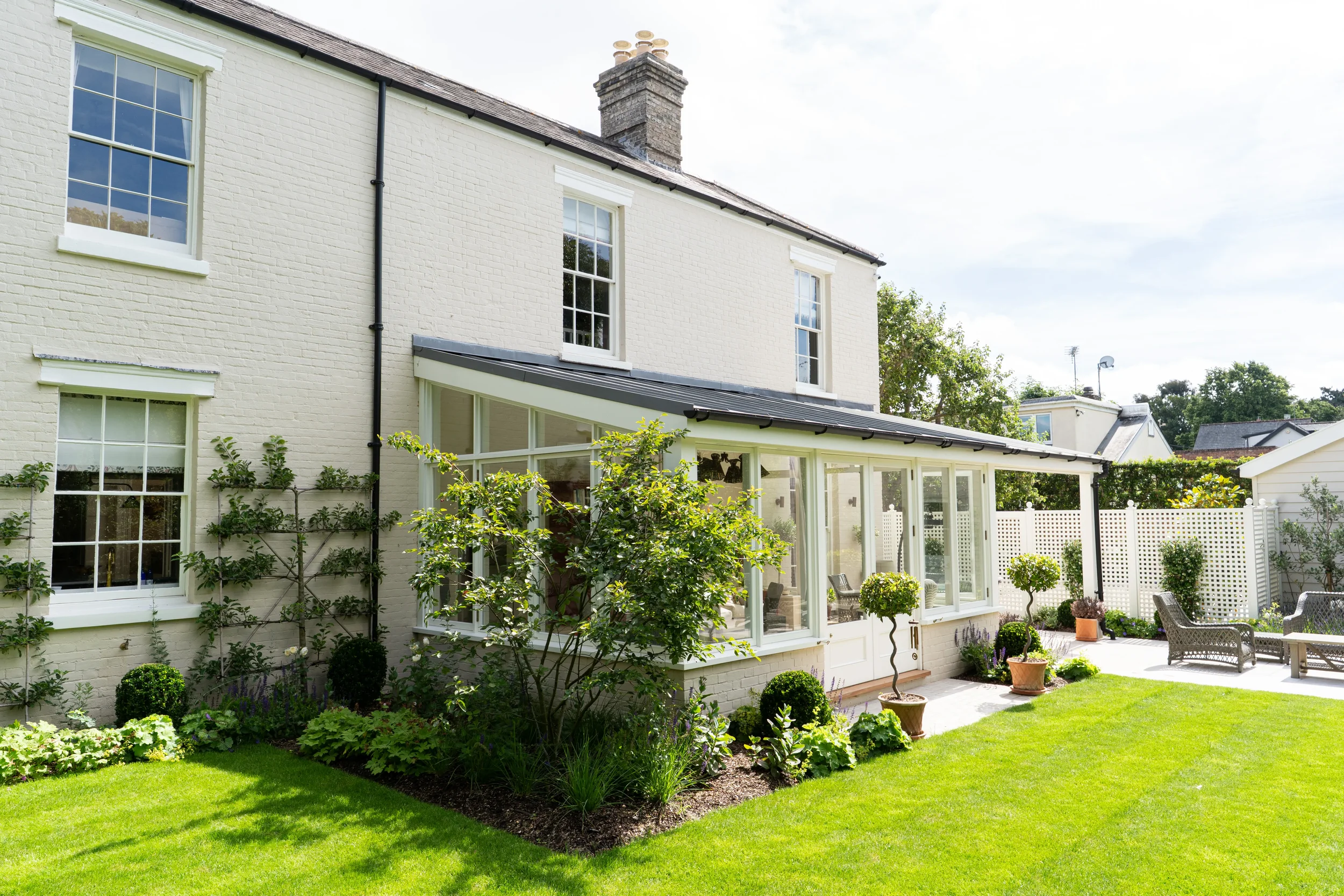 White brick house with multiple large windows and a glass-enclosed porch, surrounded by a well-maintained green garden with potted plants and trees.