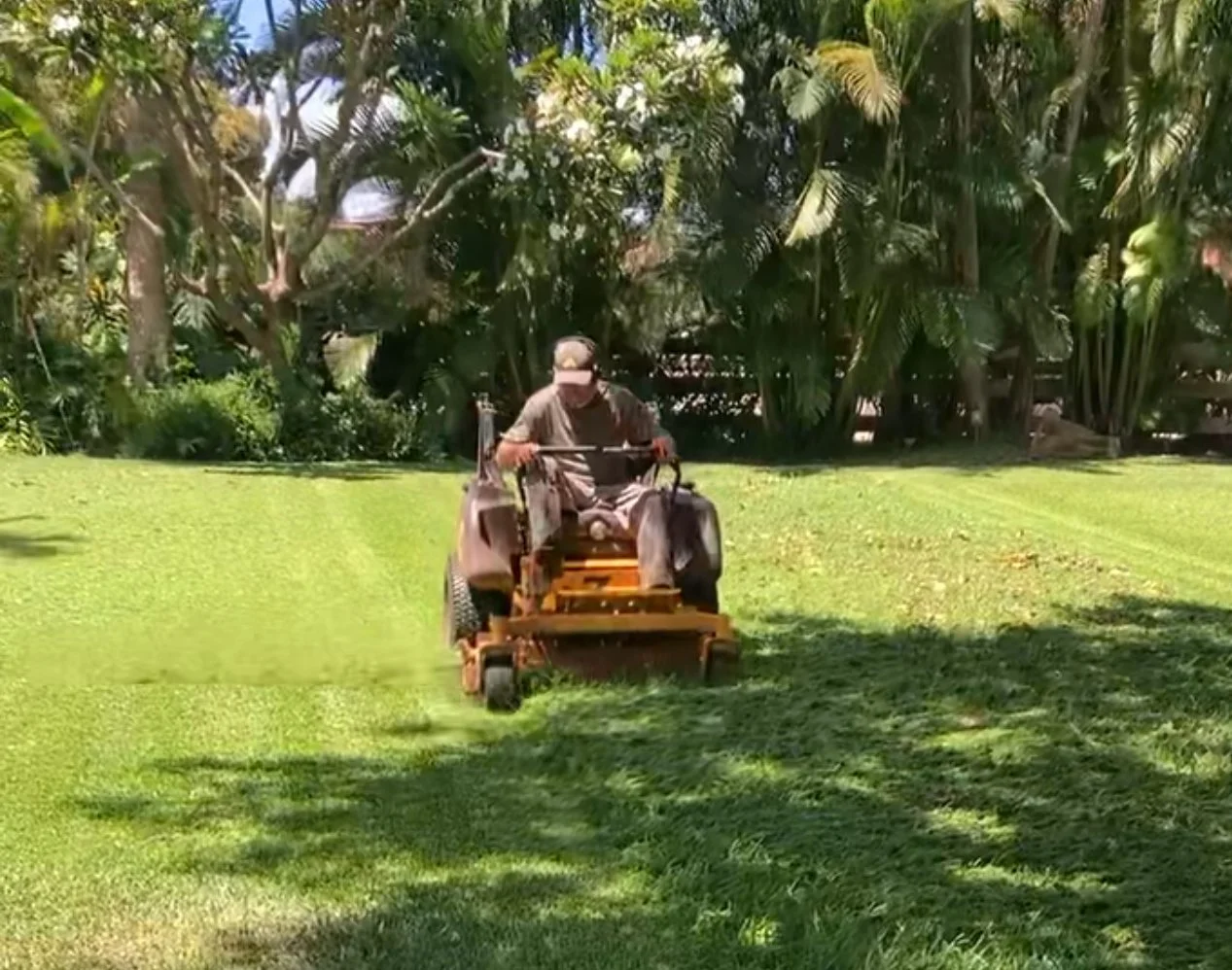 A person wearing a cap riding a lawn mower over a lush green lawn surrounded by trees and bushes.