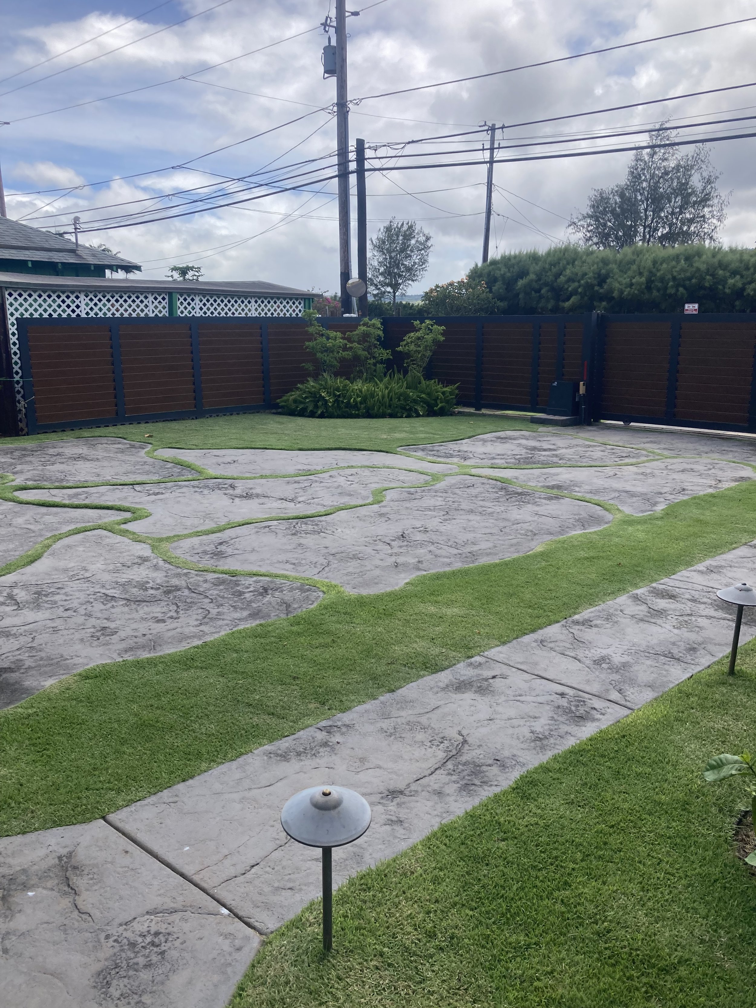 A backyard with a landscaped concrete patio featuring irregular stone patterns bordered by strips of green grass. There are two small lamp posts with domed tops visible near the patio. In the background, there is a tall wooden fence, some trees, and 