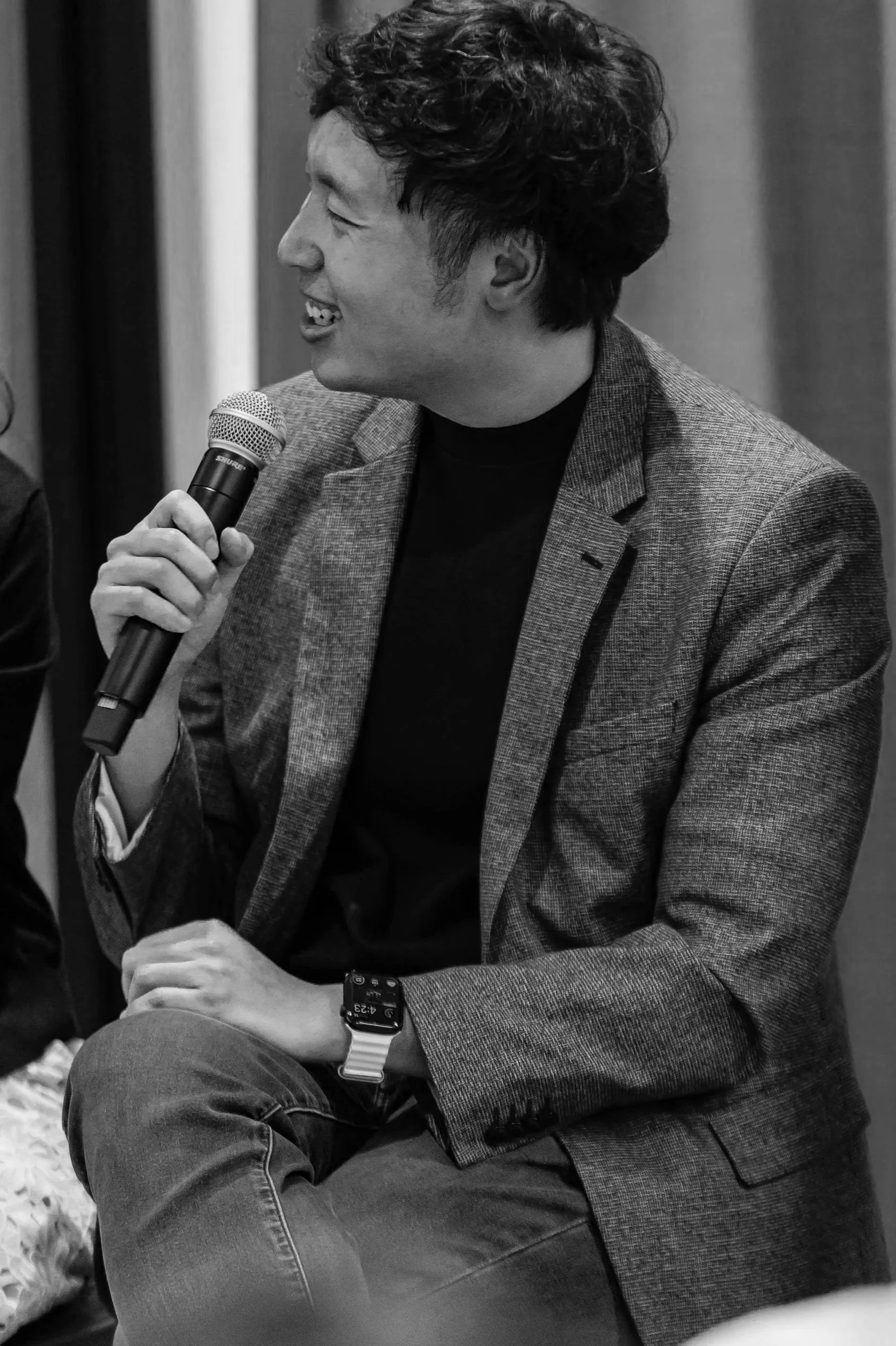 A man with curly hair, wearing a blazer and a smartwatch, smiles while holding a microphone during a discussion.