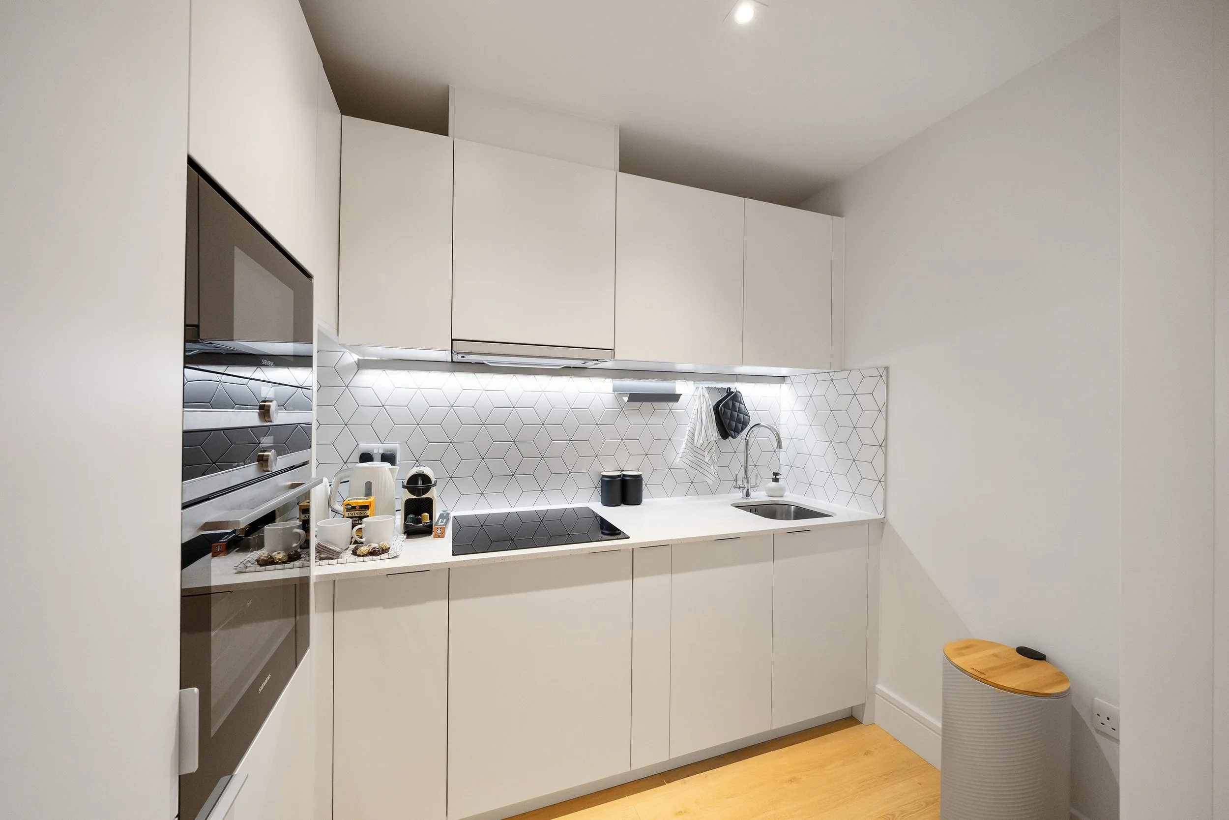 Modern white kitchen with built-in black oven, white cabinets, hexagonal tile backsplash, small kitchen appliances, dish rack, sink, and minimalist decor.