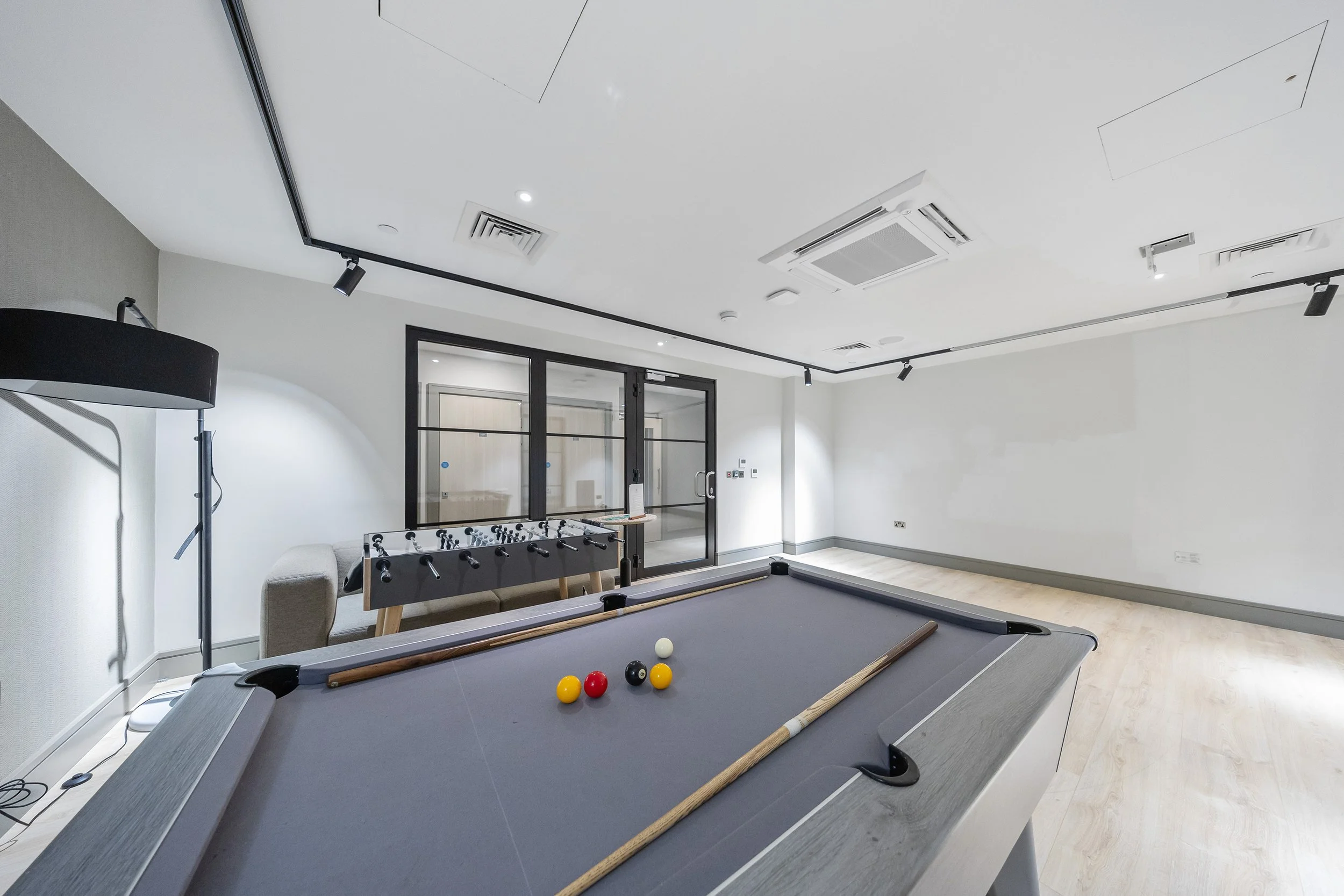 Room with a foosball table, pool table, gray sofa, black floor lamp, side table, and a glass door.