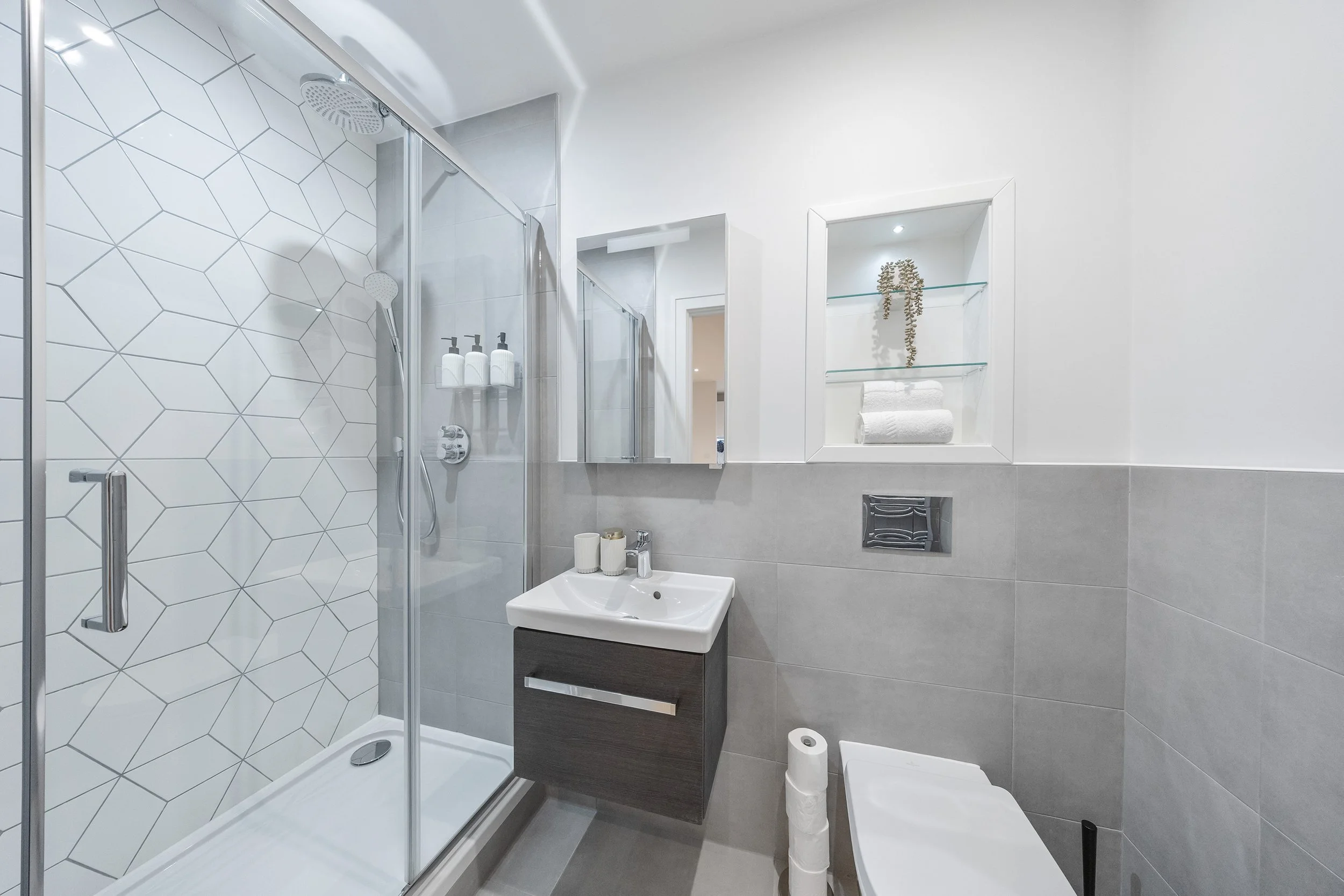 Modern small bathroom with a glass shower enclosure, white and gray tiles, a small wall-mounted sink with a mirror above, a toilet, and decorative shelving with towels and plants