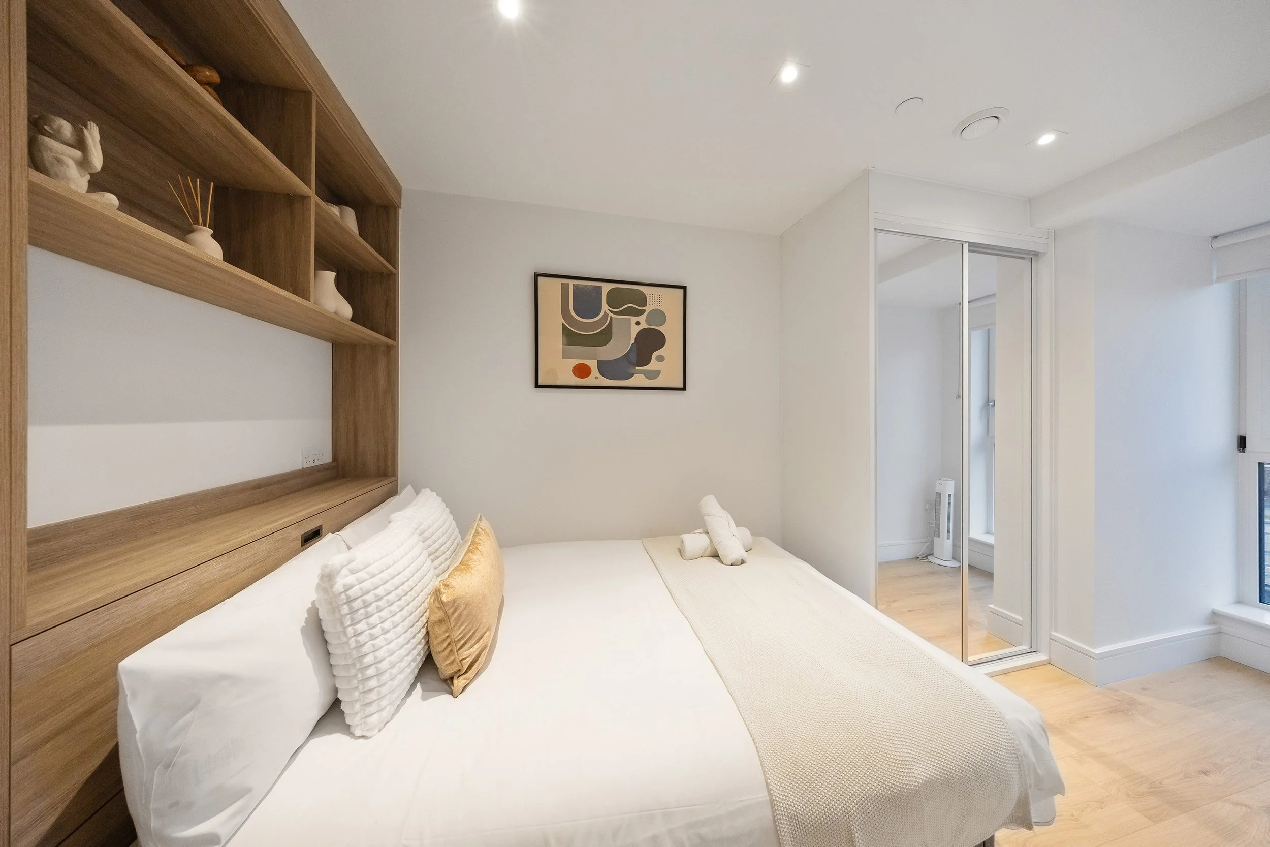 A neatly made bed with white pillows, a beige blanket at the foot of the bed, on a light-colored wooden floor, with a light wood headboard platform that has built-in shelves holding decorative items; white rolled towels are on the beige blanket.