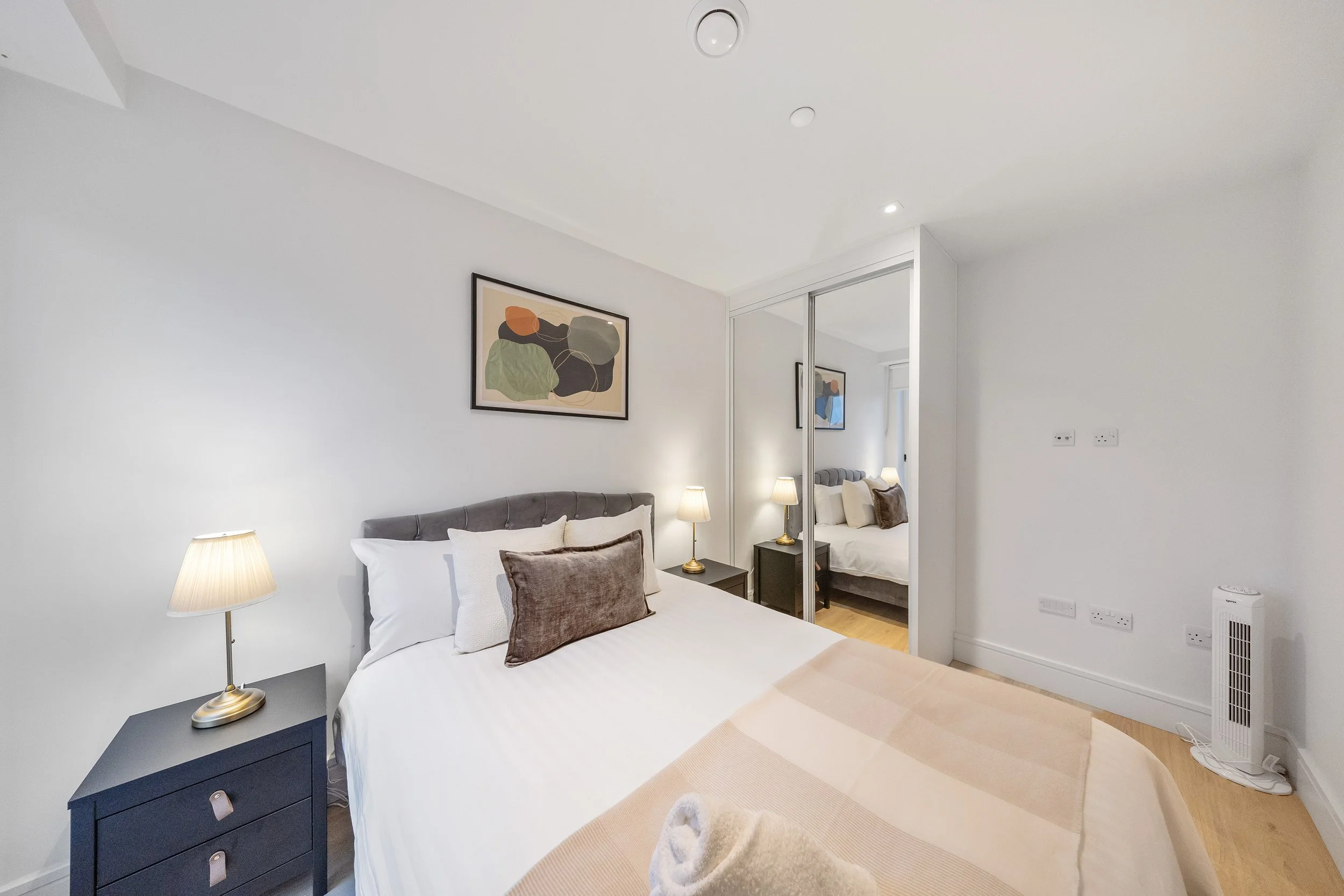 Clean modern bedroom with white walls, gray headboard, white bedding, blue throw pillows, a bedside table with a lamp, and a framed abstract artwork above the bed. There is a wardrobe with a mirror and open shelves.