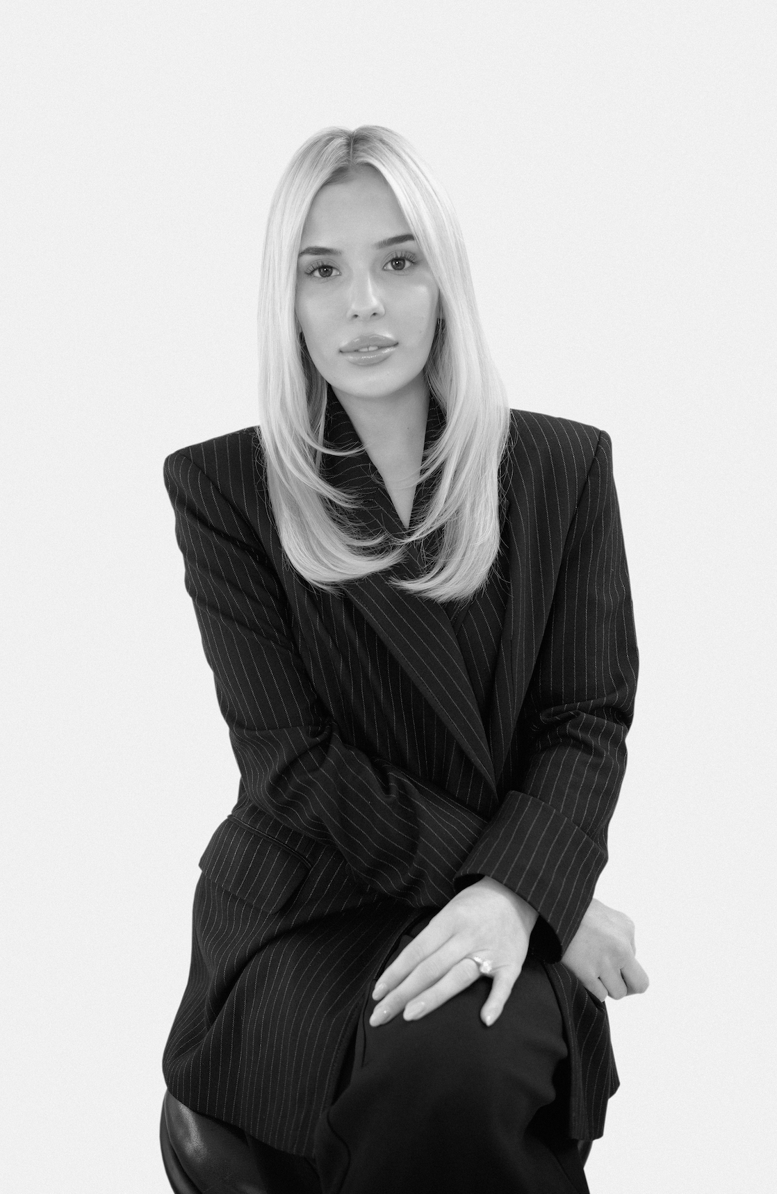 A woman with long blonde hair wearing a pinstripe suit sitting against a plain background.