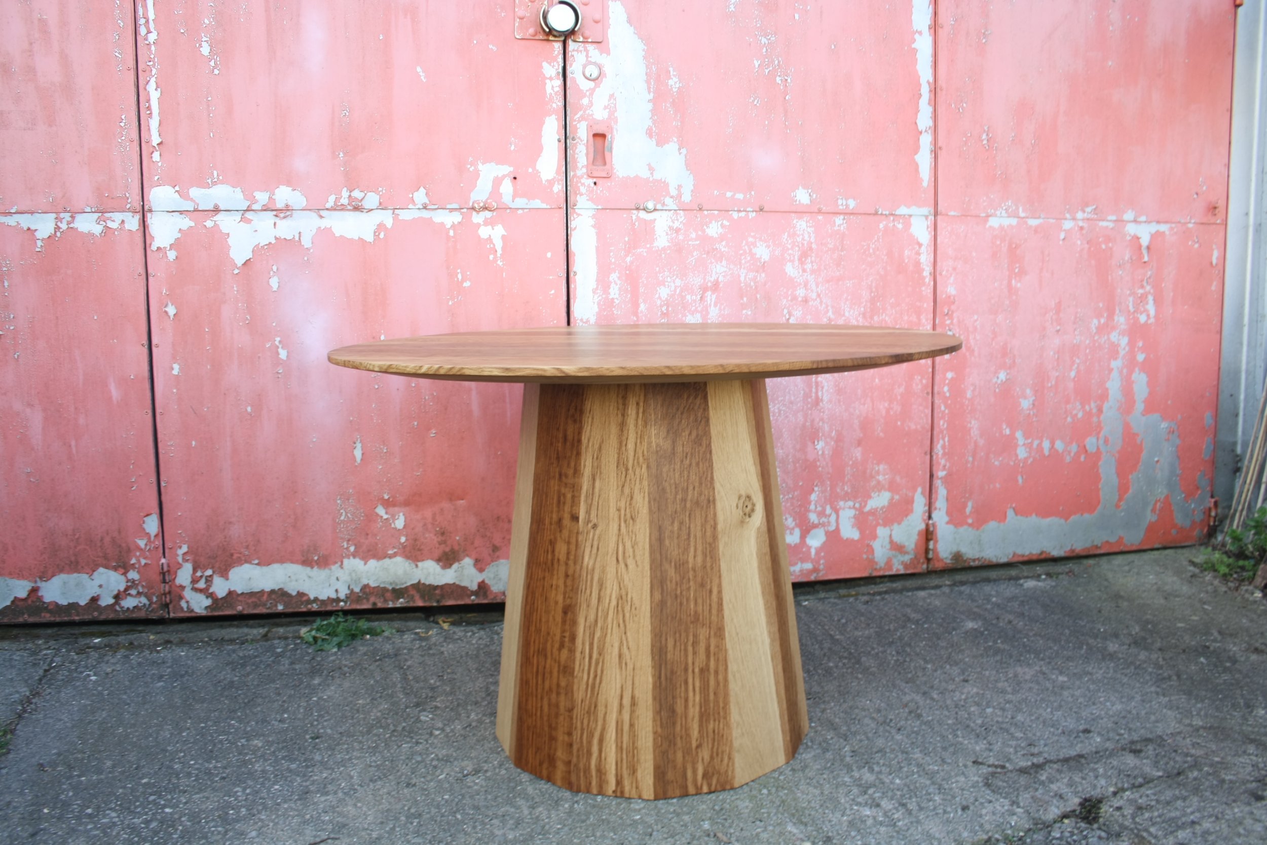 A round wooden table with a tapered, faceted base is placed outside in front of a weathered pink metal wall with peeling paint.
