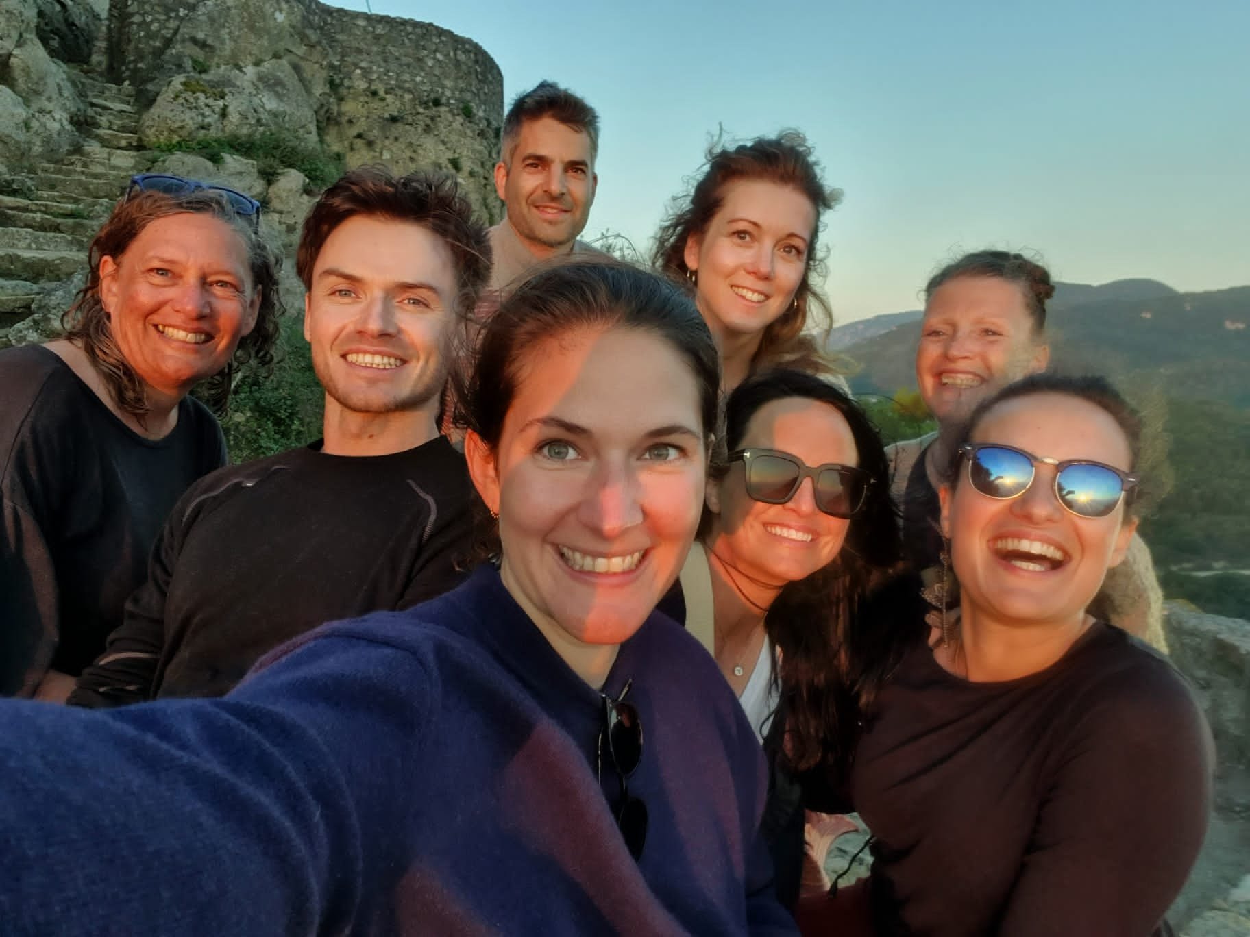 A group of ten people smiling and posing for a selfie outdoors during sunset, with mountains and a stone staircase in the background.