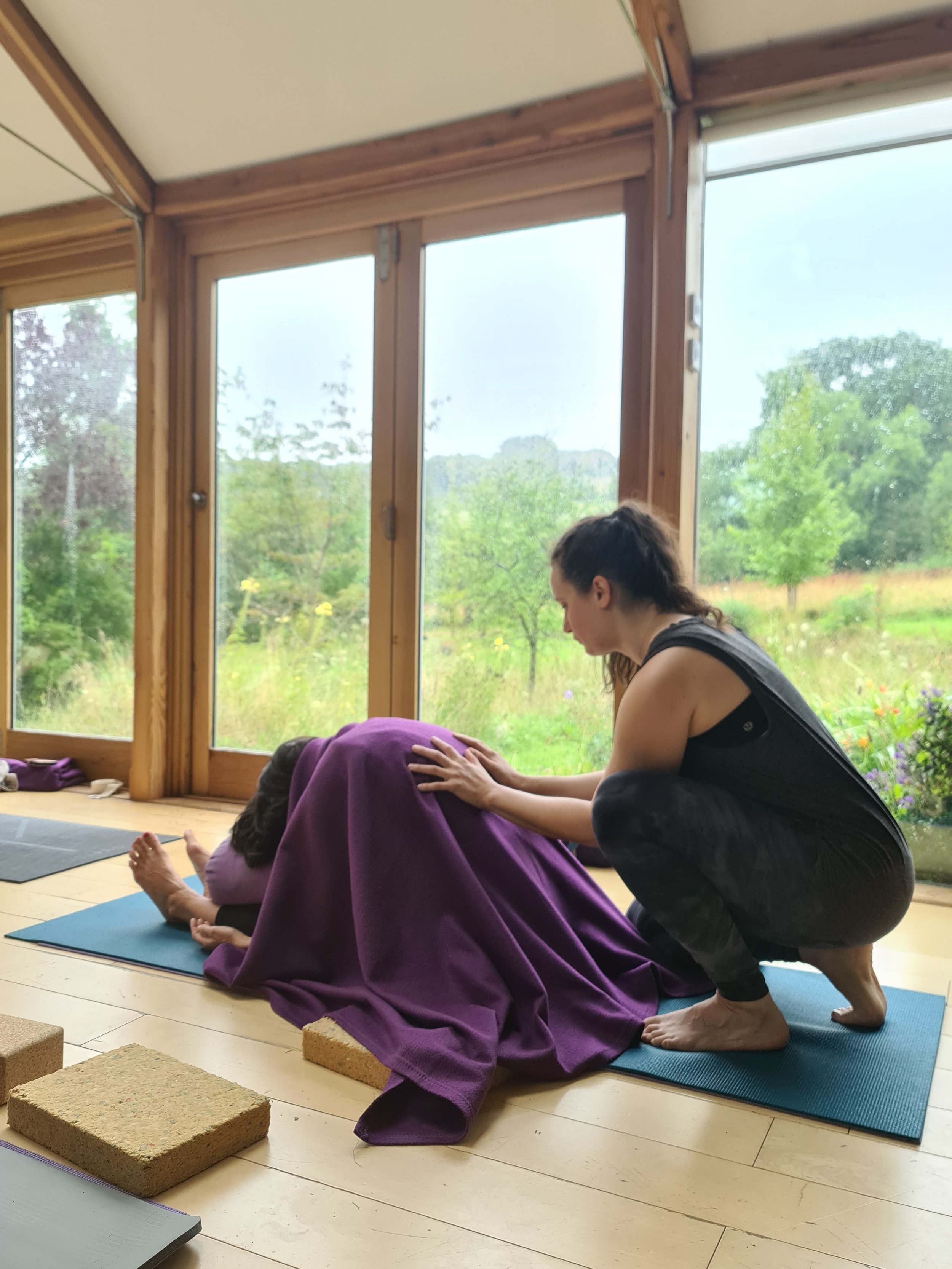 A woman practicing yoga with a young girl, who is covered with a purple blanket, on a blue yoga mat in a room with large windows showing a green outdoor landscape.