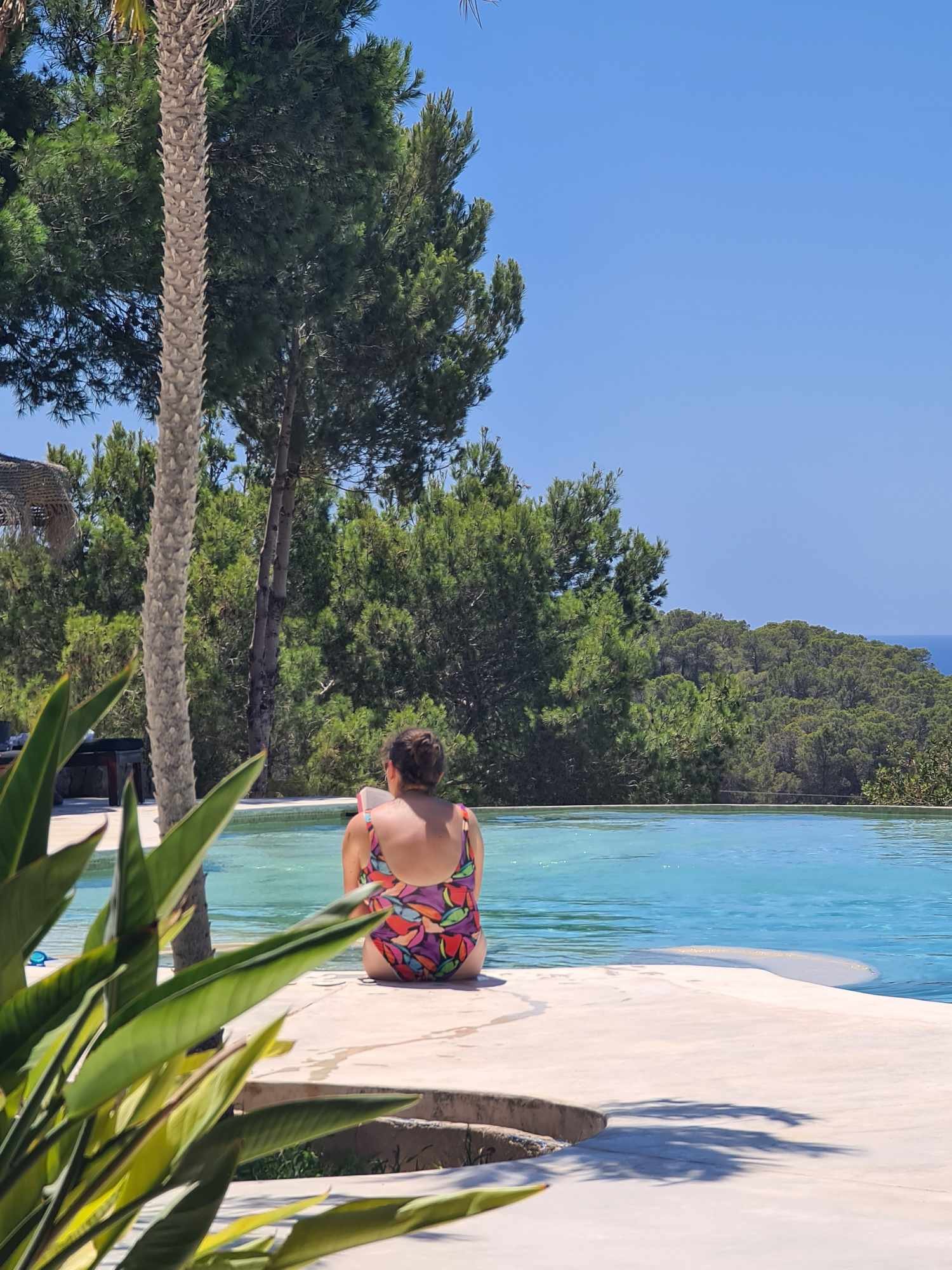 A person in a colorful swimsuit sitting on the edge of a pool, surrounded by trees and bushes with a clear blue sky above.