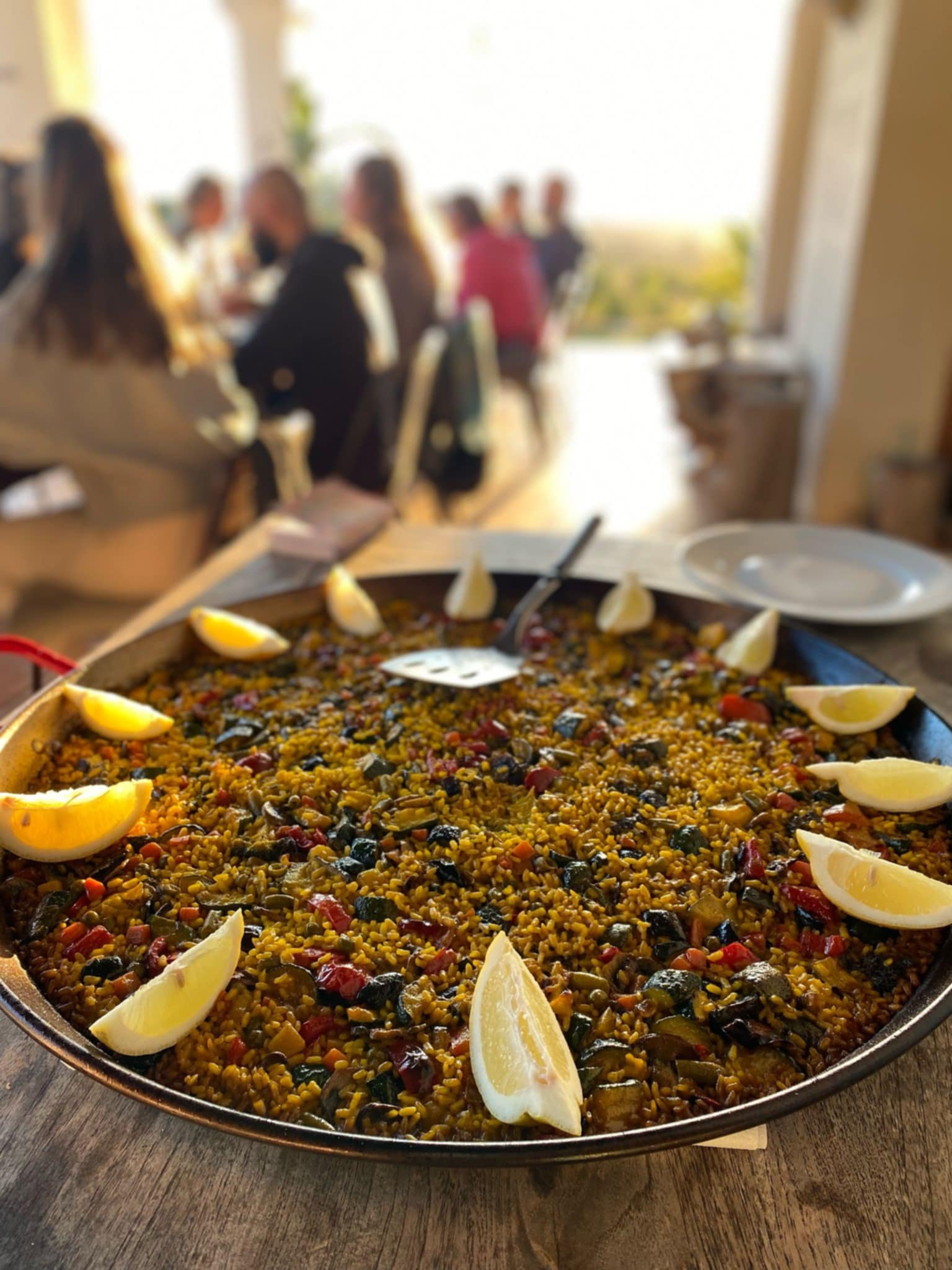 Large serving tray of seasoned rice with vegetables, garnished with lemon wedges on a wooden table in a bright restaurant setting with blurred diners in the background.