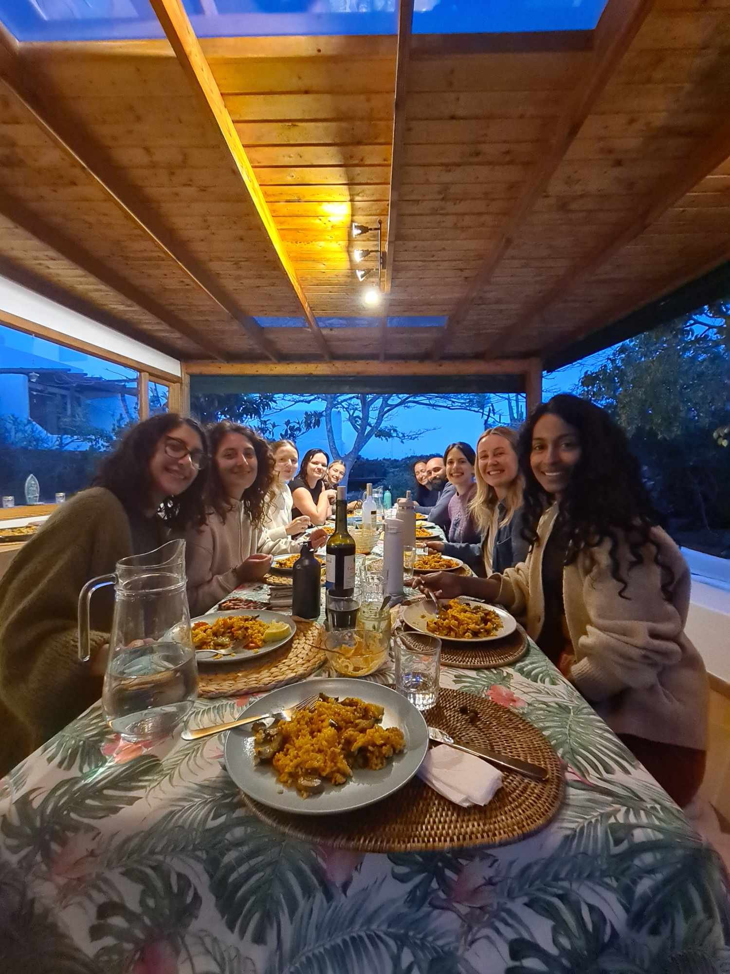 A group of people sitting around a long dining table enjoying a meal together in a cozy, wood-paneled room with large windows showing an outdoor scene at dusk.