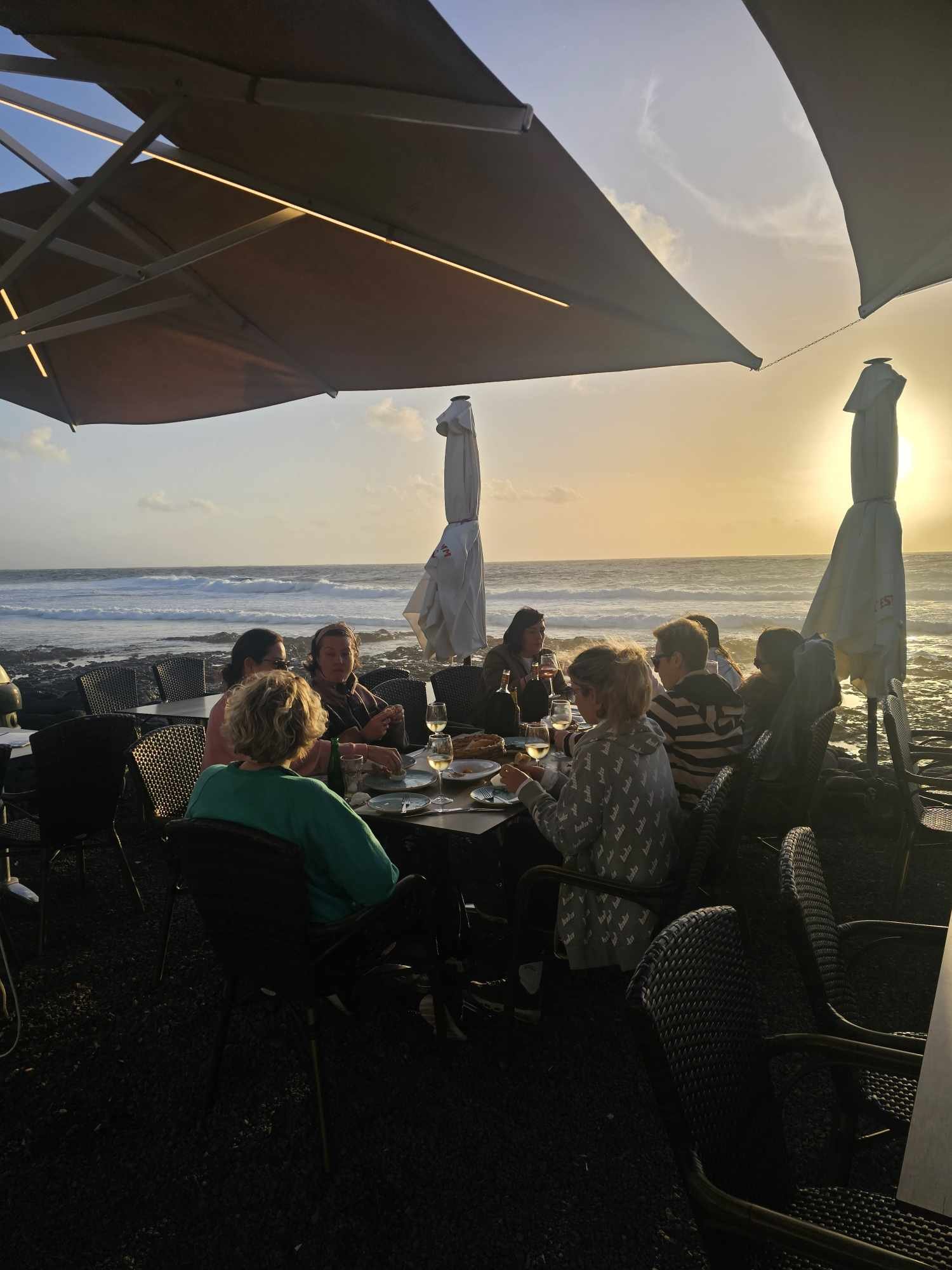 People dining at an outdoor restaurant near the beach at sunset, with large umbrellas and ocean in the background.