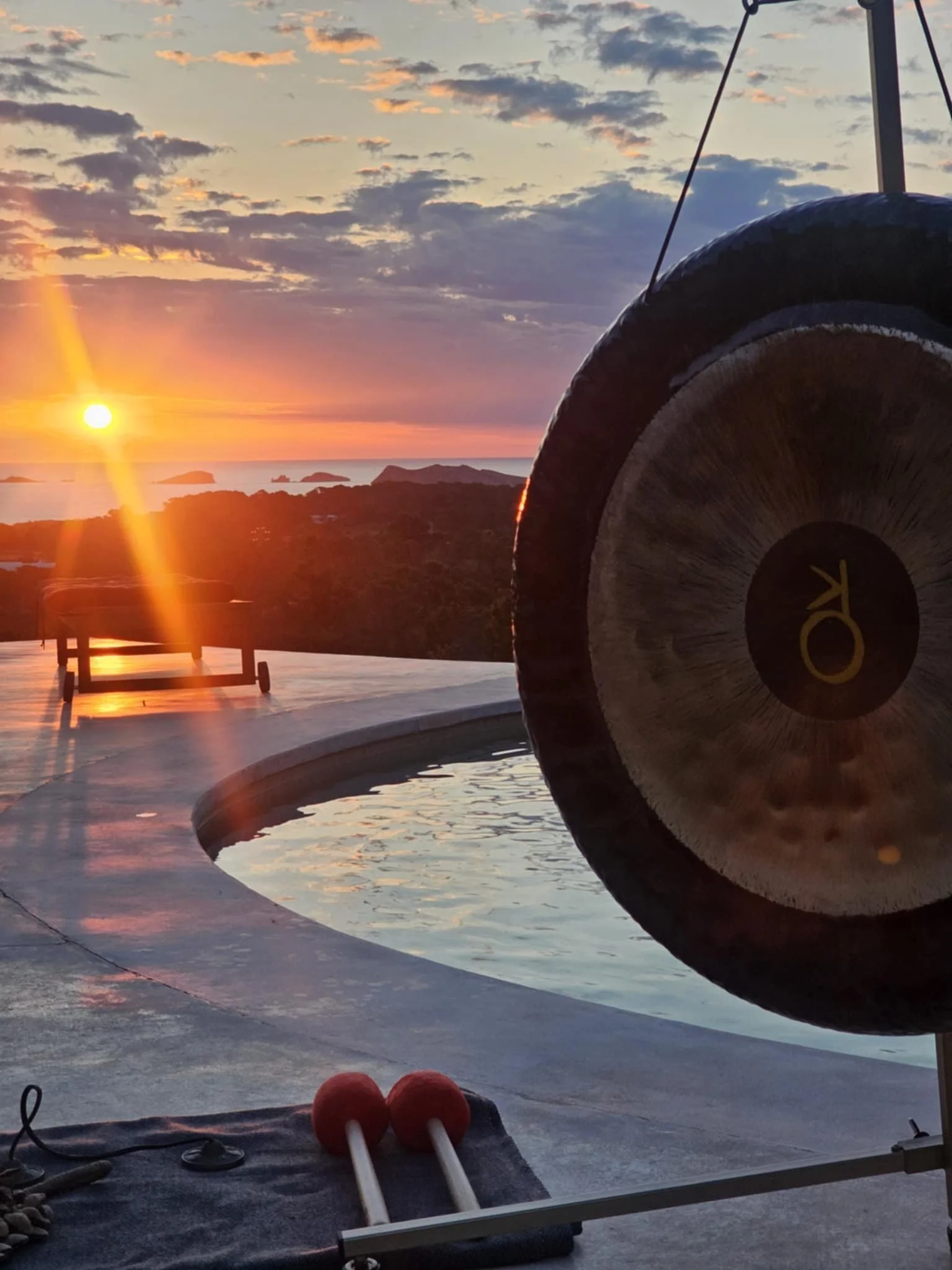 A scenic sunset view over the ocean with a wellness setup including a large gong, a bench, and a massage technique tool with red balls, near a pool.