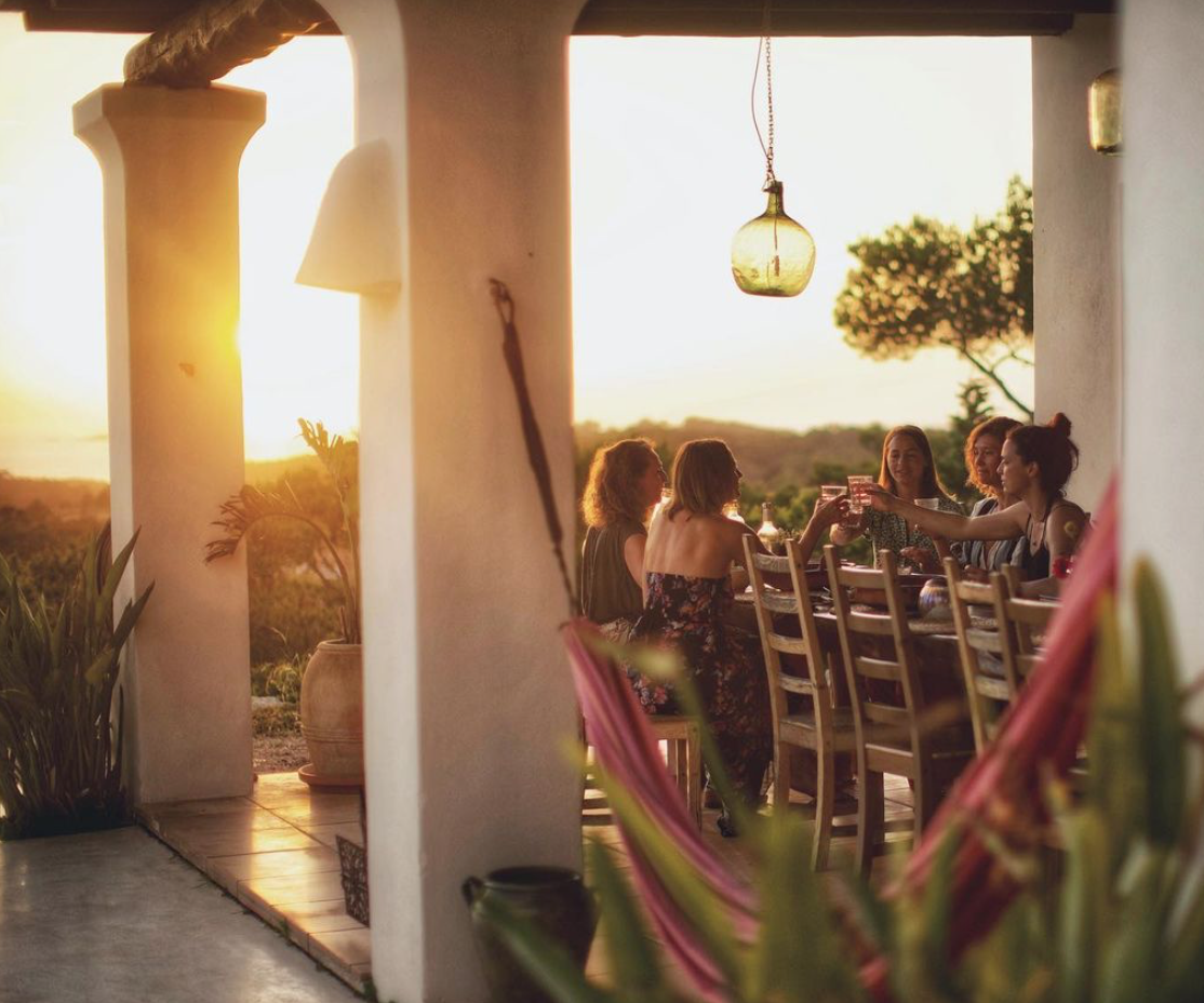 Group of six women sitting at a dining table outdoors during sunset, enjoying drinks and conversation, with a scenic natural landscape in the background.