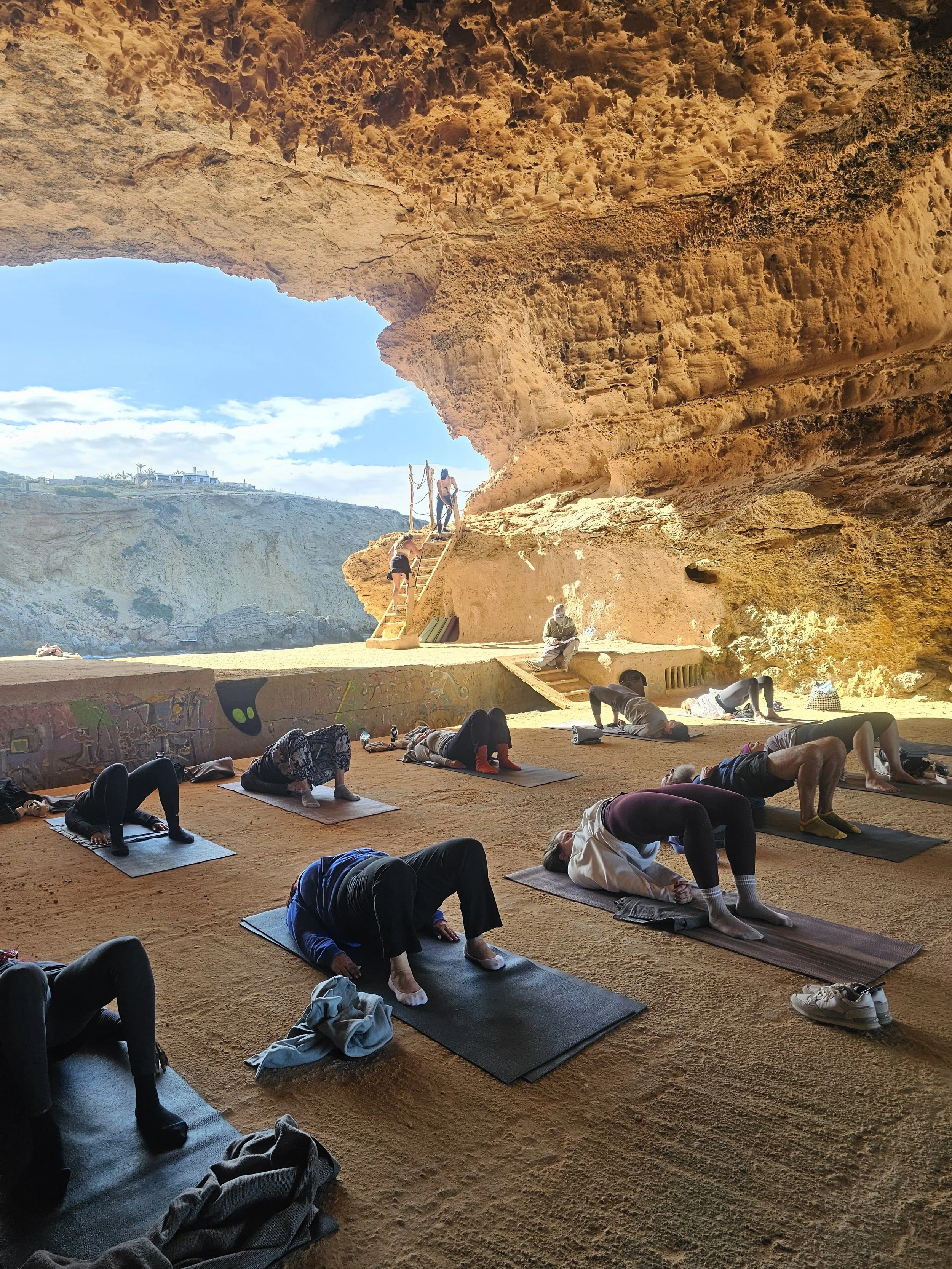 People practicing yoga in a shaded cave with sandy floor and large rock formations, overlooking a coastline with blue sky and clouds.