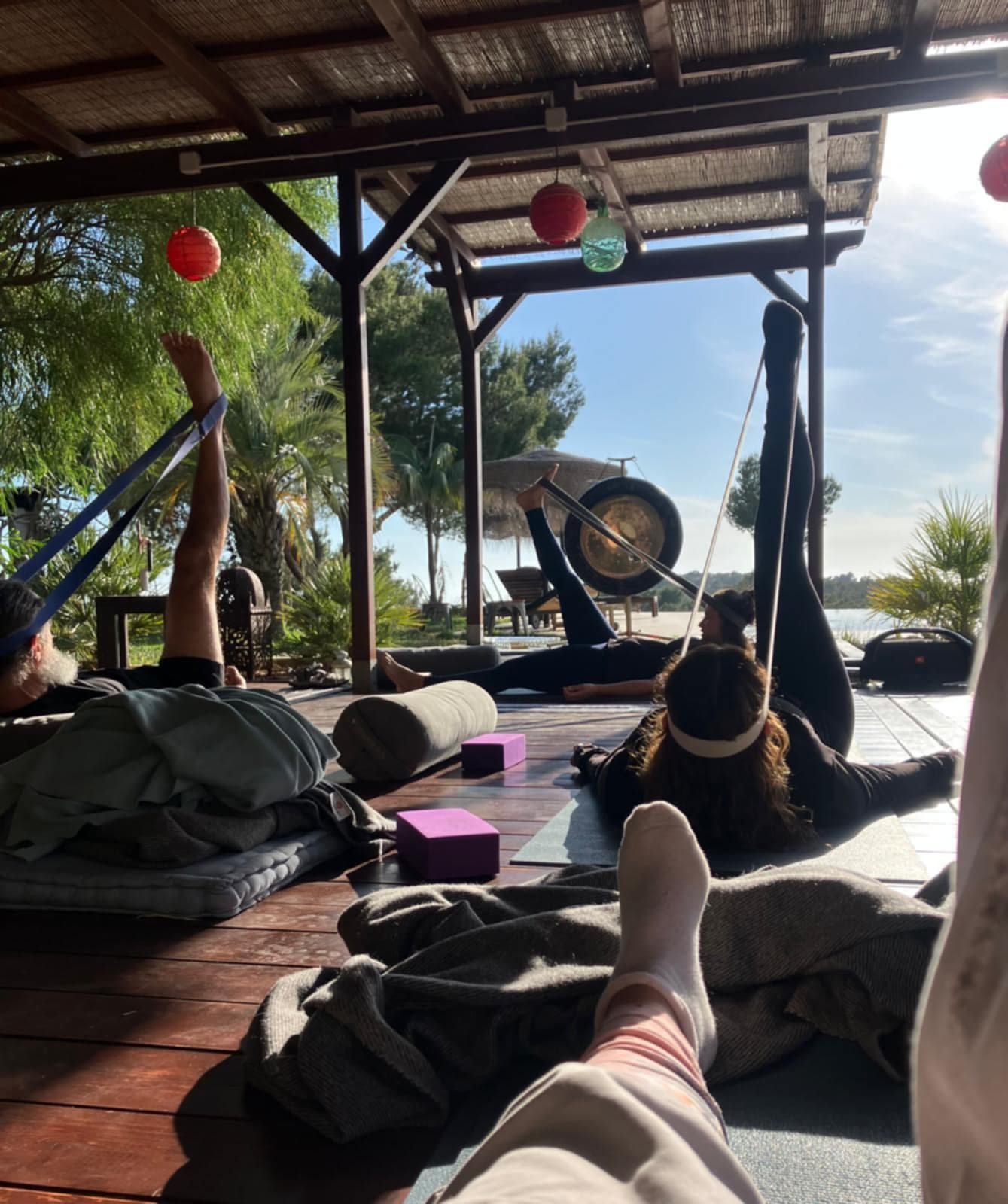 People practicing yoga outdoors on a wooden deck near a lake, with some using resistance bands and a gong, surrounded by trees and colorful paper lanterns.