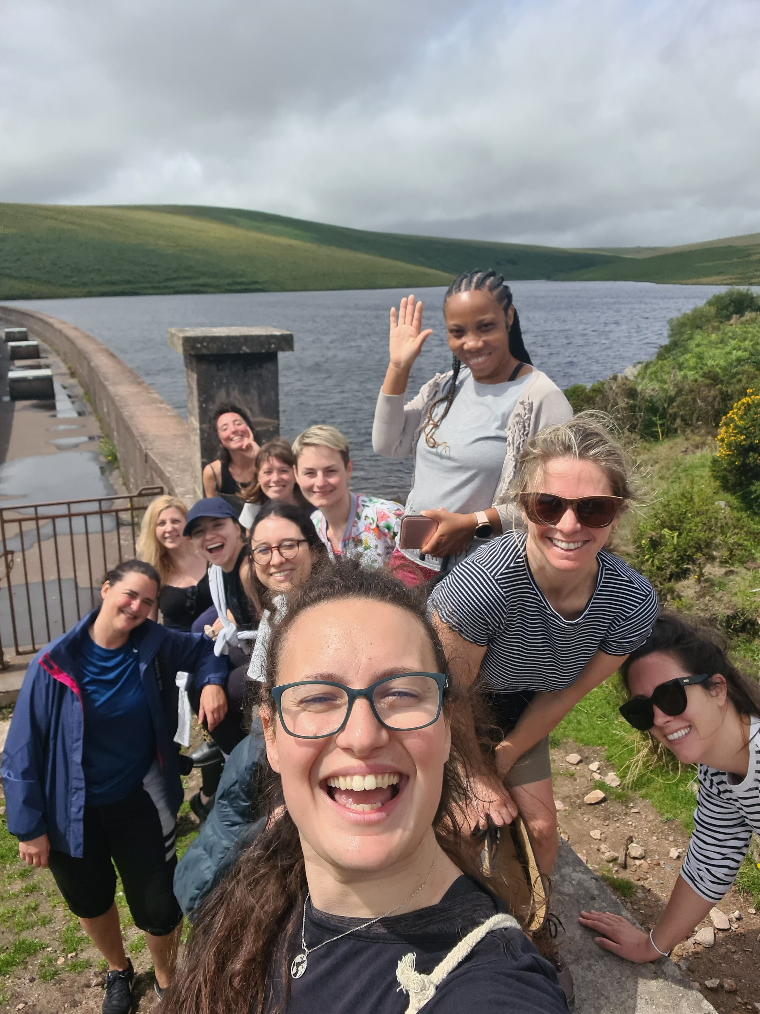 Group of smiling women taking a selfie outdoors near a lake with green hills in the background.