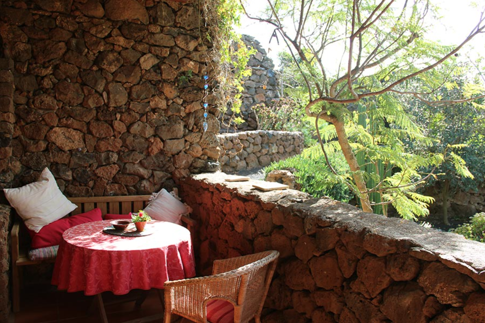 Outdoor patio with a red round tablecloth, two pillows, a small plant, and a fruit bowl, surrounded by a stone wall and lush greenery.