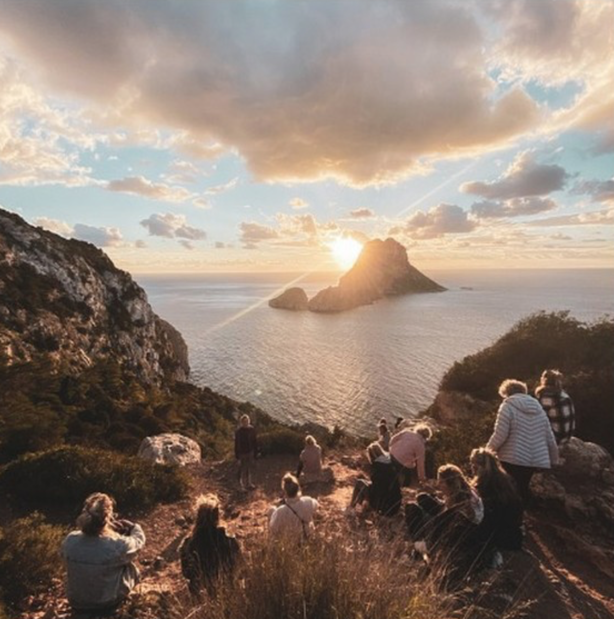 People watching sunset over the ocean from a rocky hillside with rock formations in the water.
