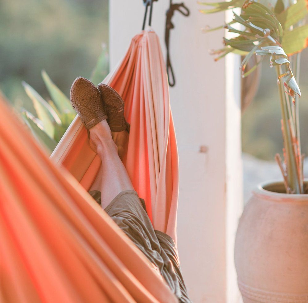 Person relaxing in an orange hammock on a porch, wearing brown slippers and khaki shorts, with plants in a large pot nearby.