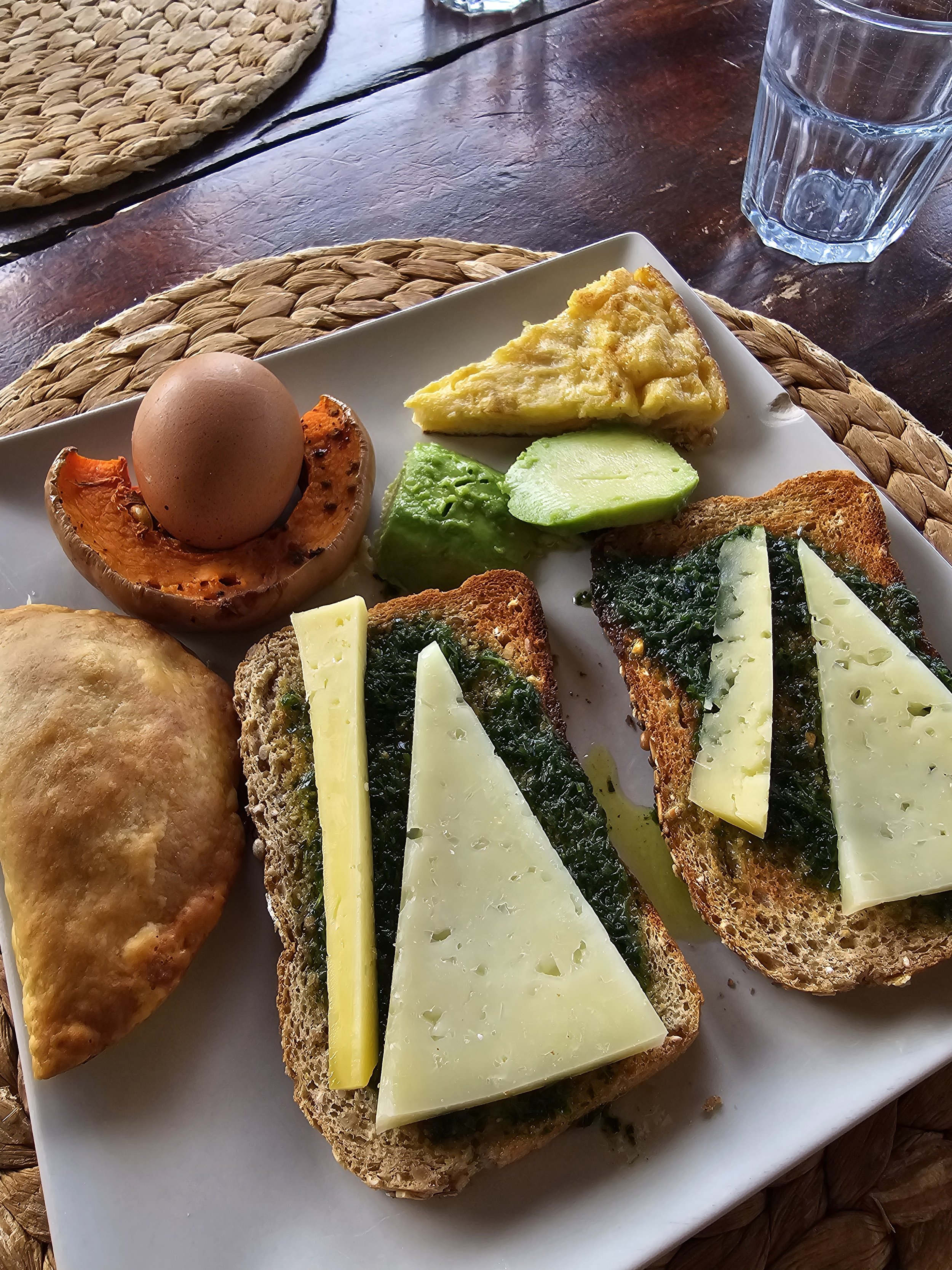 A white plate with two slices of toast topped with spinach and cheese, a baked empanada, a hard-boiled egg in an avocado half, a slice of quiche, and avocado slices. A glass of water is nearby on a wooden table with a woven placemat.