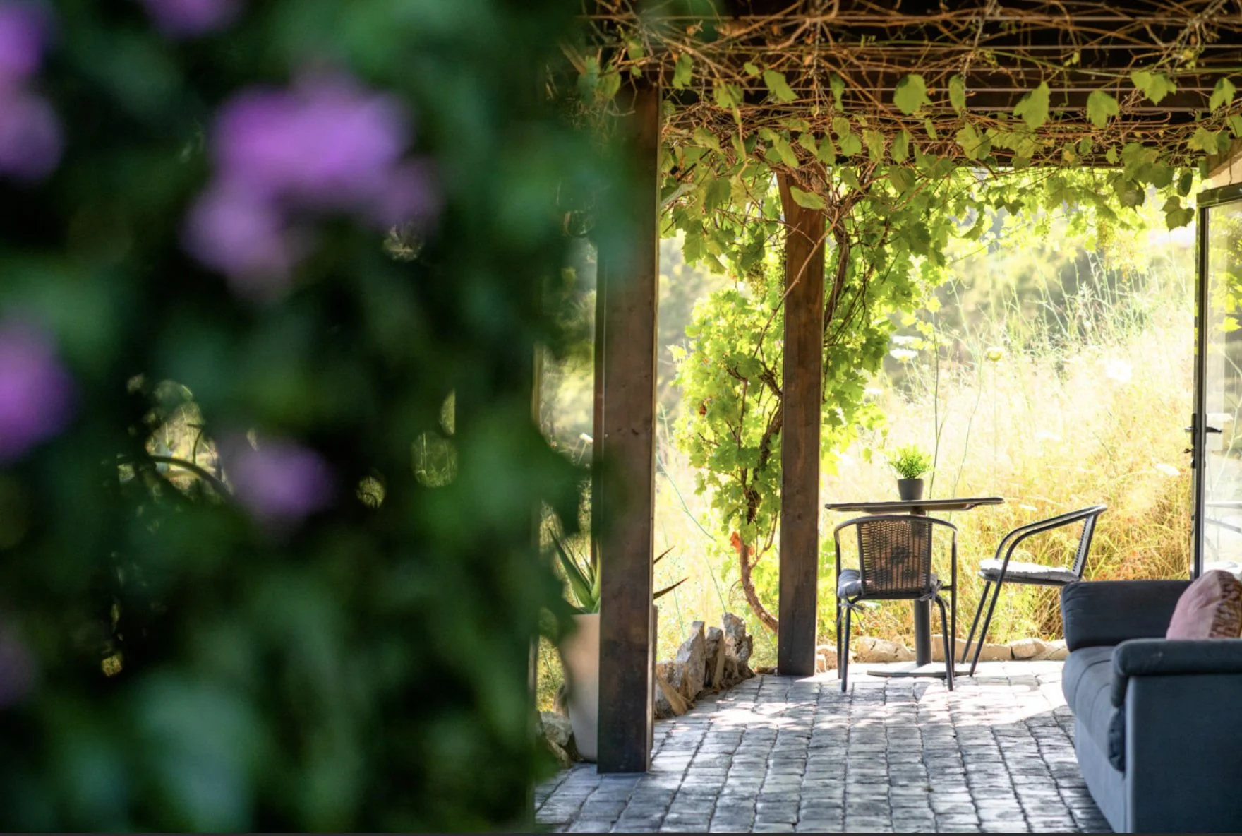 A cozy outdoor patio with a stone floor, surrounded by lush greenery and vines, featuring a small table with a plant and two chairs, one of which is a wicker chair, outside a house or cafe.