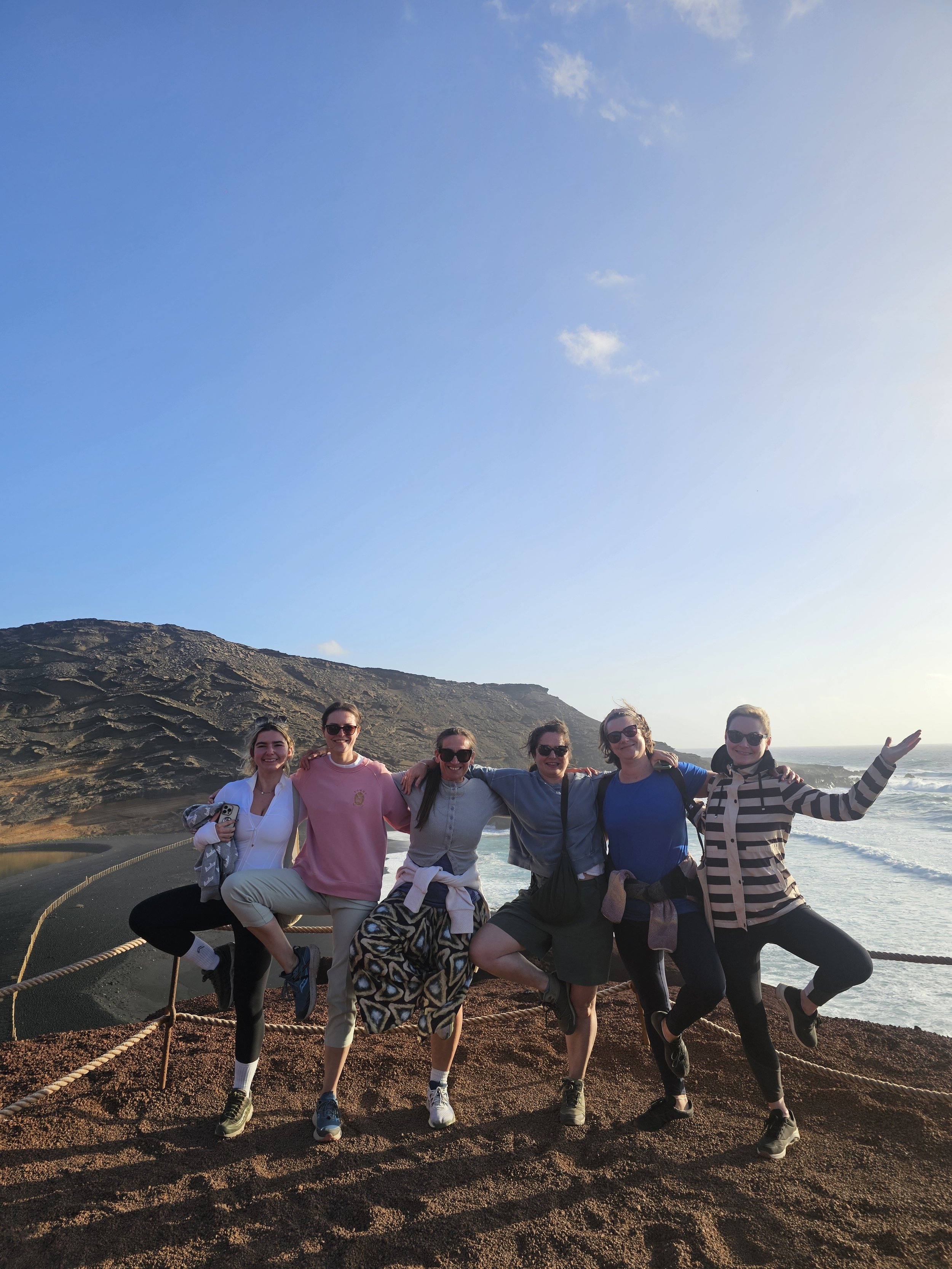 Group of six women standing on a sandy hill near the ocean, smiling, with a cliff and blue sky in the background, posing with arms around each other.