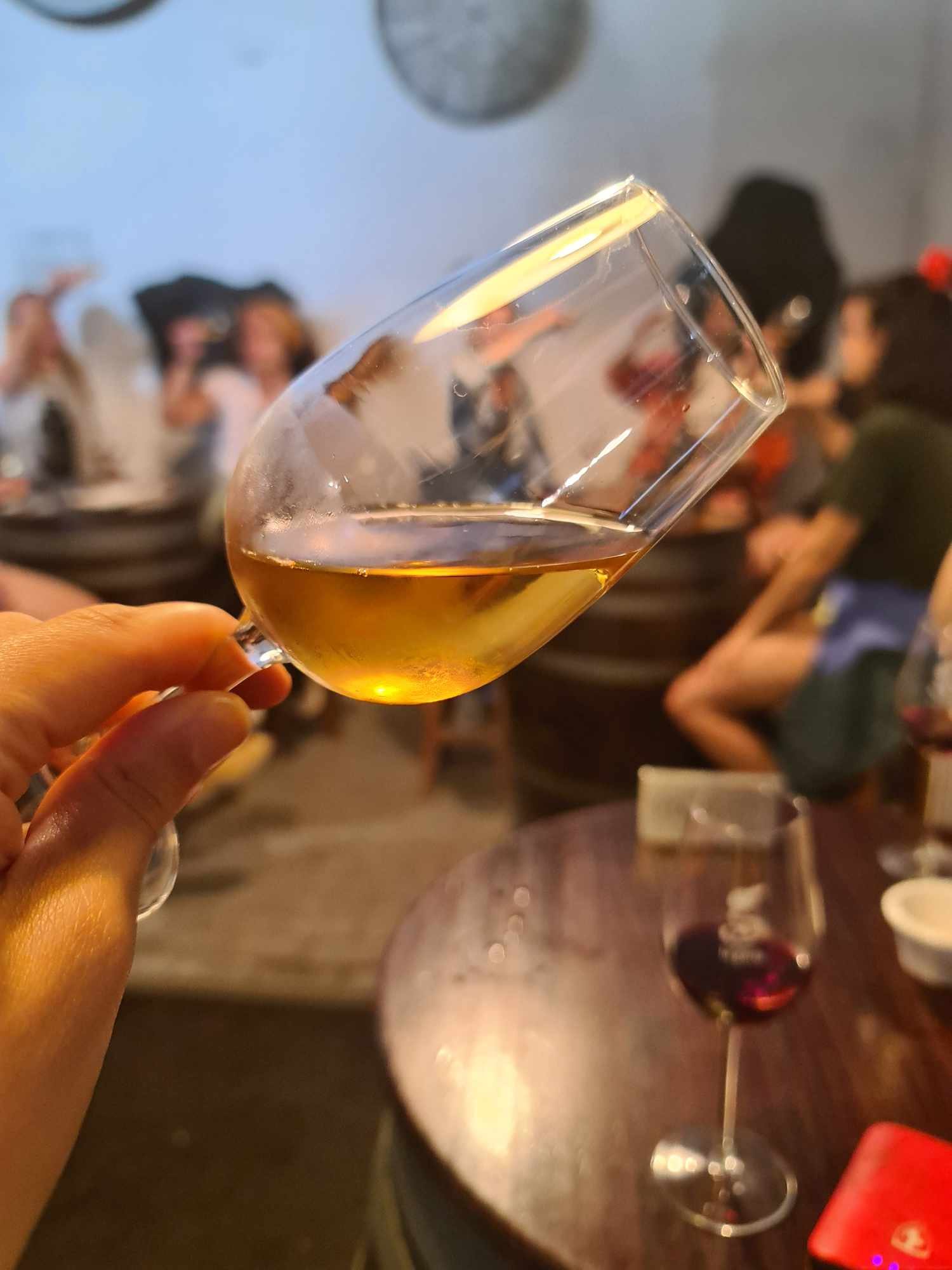 A hand holding an upturned wine glass with a golden beverage, with a blurred background of people socializing at a party or gathering.