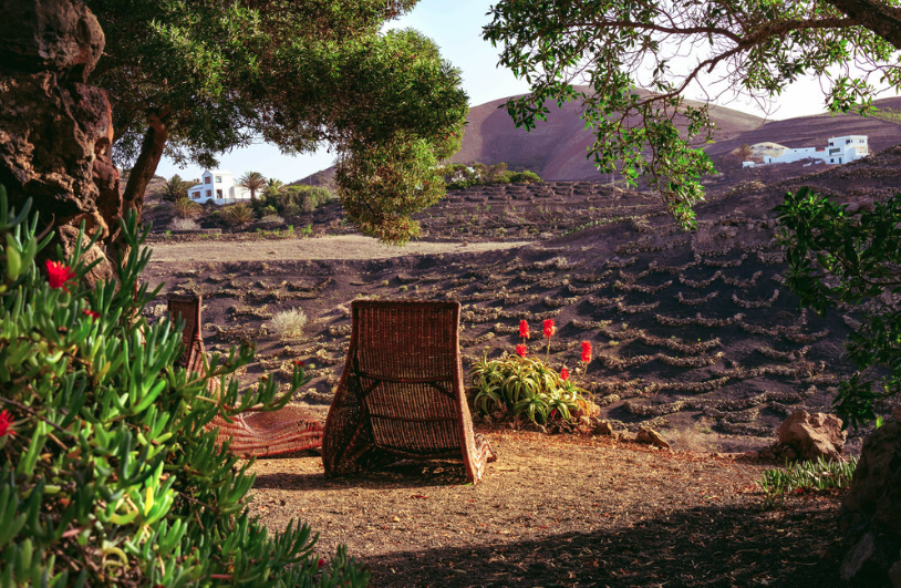 A wicker outdoor chair facing a terraced hillside with sparse vegetation in a sunny, arid landscape, with white buildings visible in the distance under a clear sky.