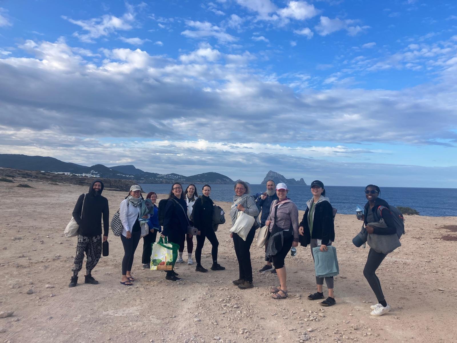 Group of eleven people standing on a rocky beach with mountains and the ocean in the background, under a partly cloudy sky.
