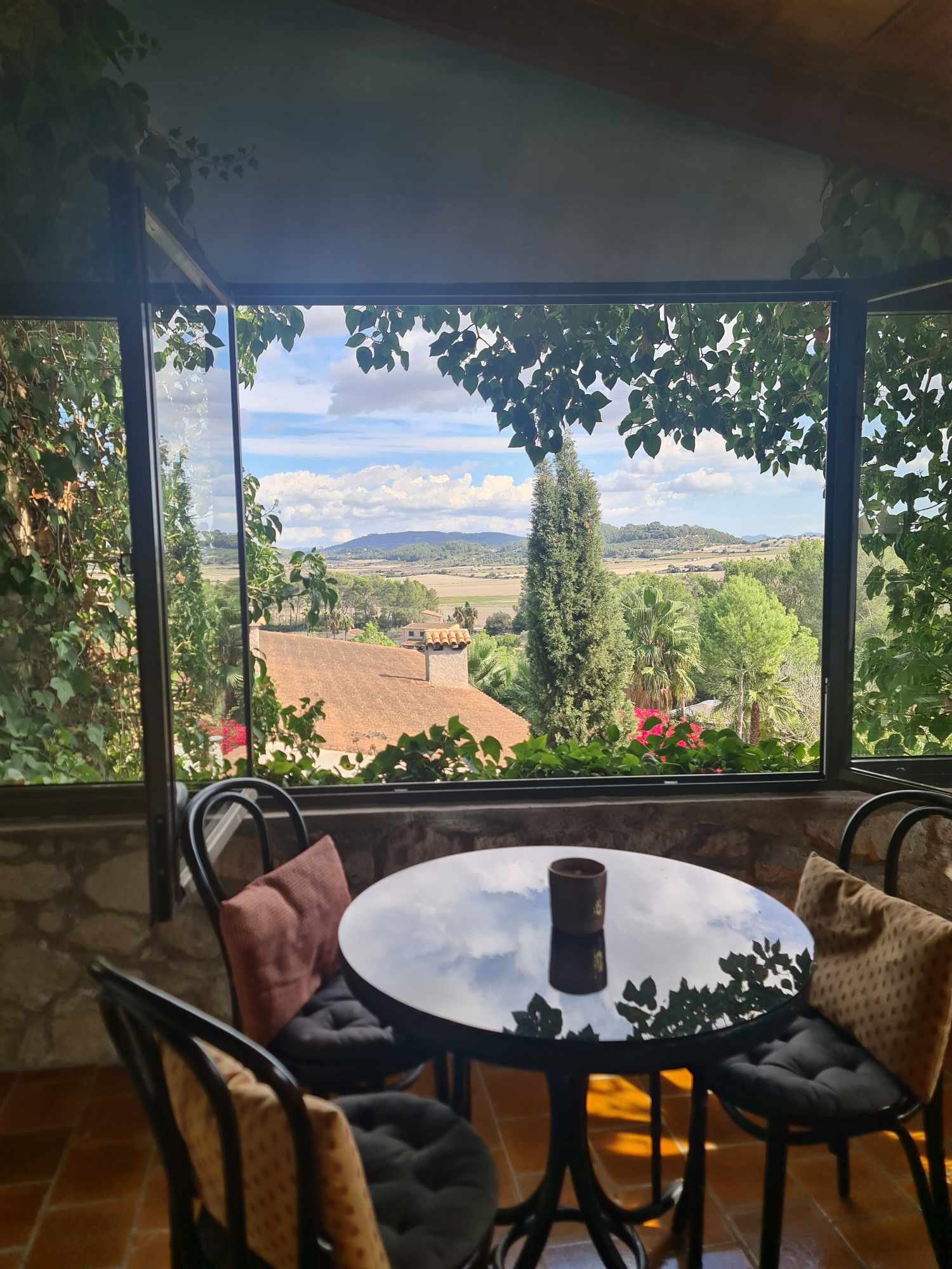 Indoor dining area with a round glass table and four chairs, featuring view through large open window of a lush green landscape with trees, a roof, and distant hills under a partly cloudy sky.