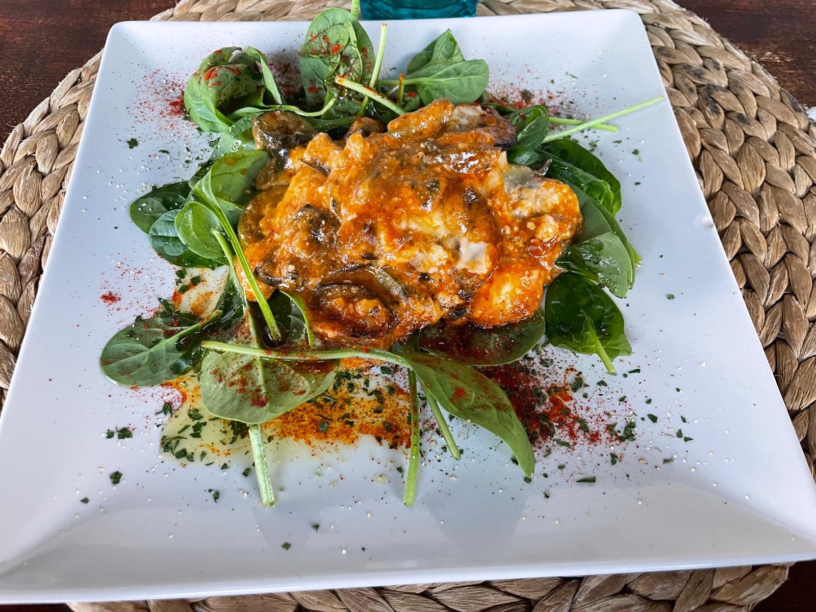 A plate of baked dish topped with cheese, served over a bed of fresh greens and leafy herbs, garnished with dried herbs and paprika on a white square plate.