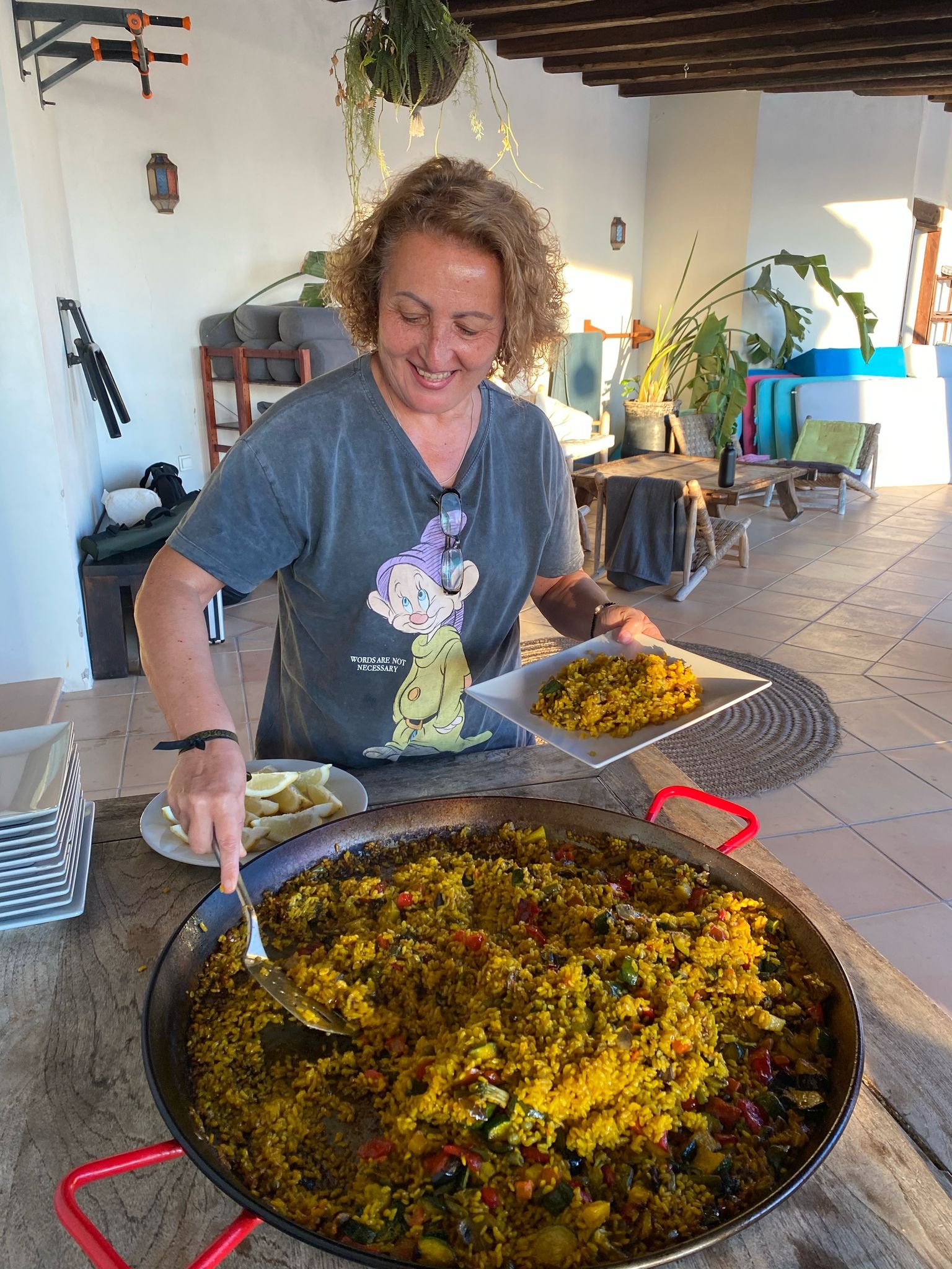 A woman with curly blonde hair smiles as she serves a large pan of yellow rice with vegetables onto a plate. The setting is a bright, cozy room with wooden beams, plants, and casual furniture.