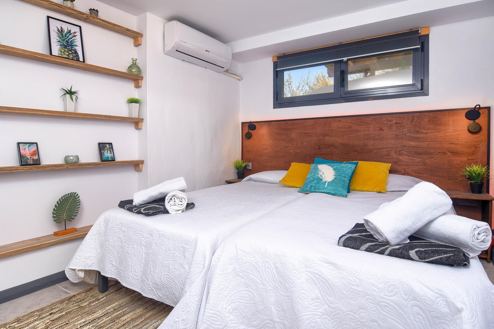 A bedroom with two beds, white quilted bedspreads, and colorful pillows including yellow and teal, in front of a wooden headboard. There are rolled towels on the beds. The room features a wall-mounted air conditioner, a window with black framing, and floating wooden shelves decorated with plants, framed pictures, and decorative objects.