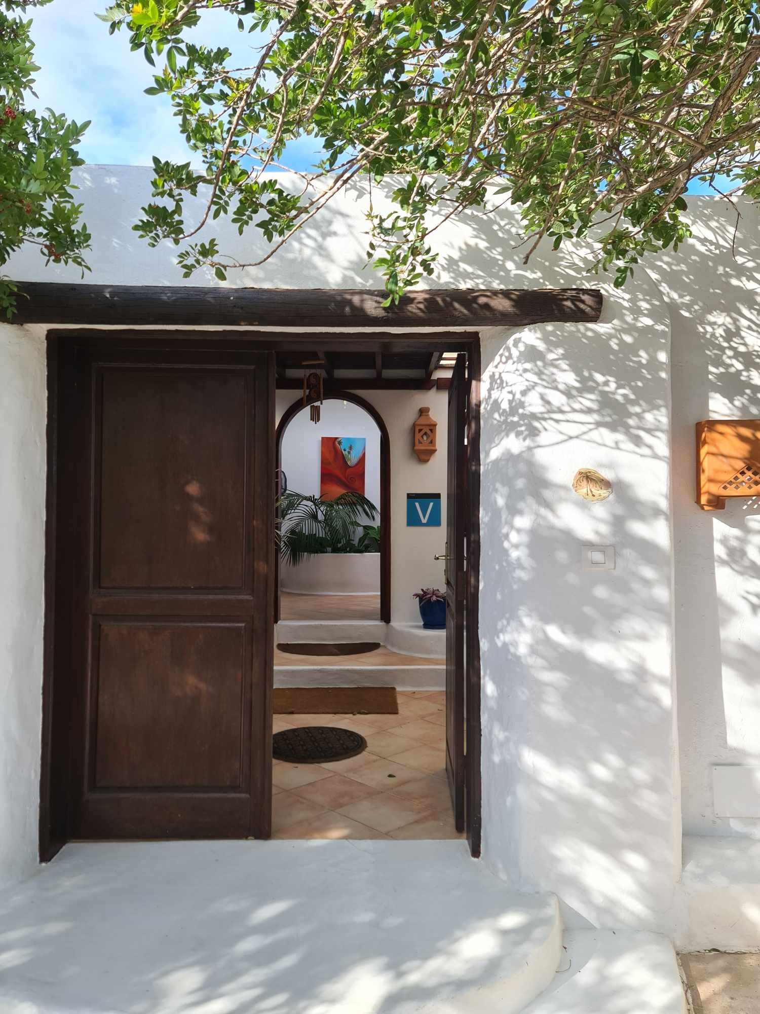 Open wooden door leading into a white stucco building with Mediterranean decor, interior includes lush green plants, a colorful painting, a blue 'V' sign, and patterned rugs, with sunlight filtering through nearby trees casting shadows.