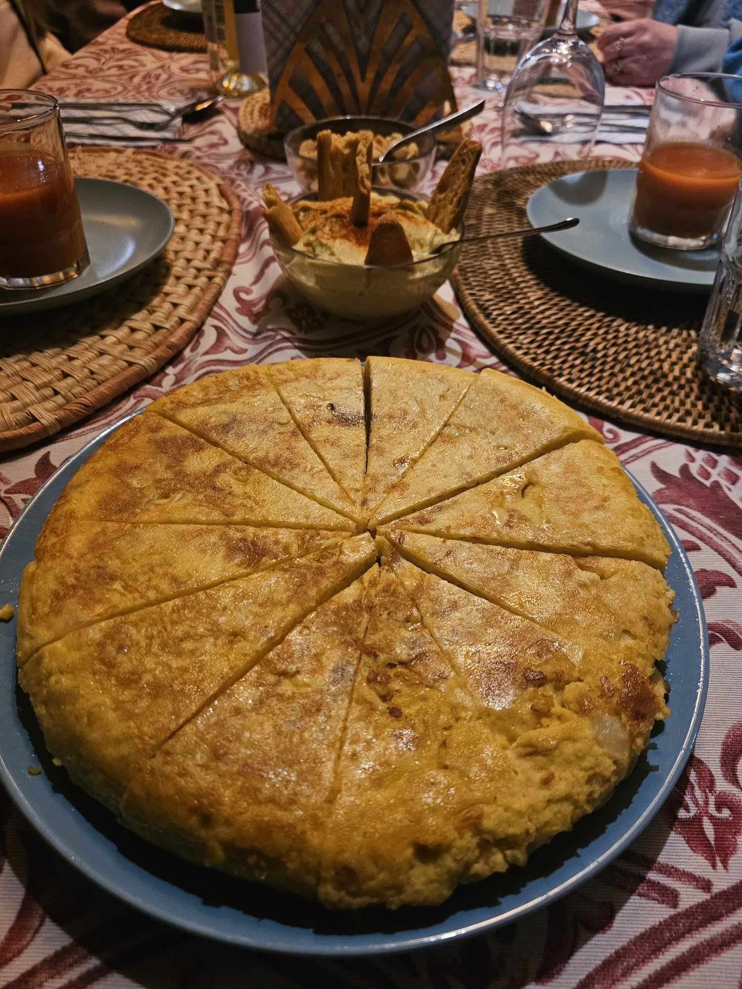 A large Spanish tortilla on a blue plate on a dinner table surrounded by drinks, bowls, and place settings.