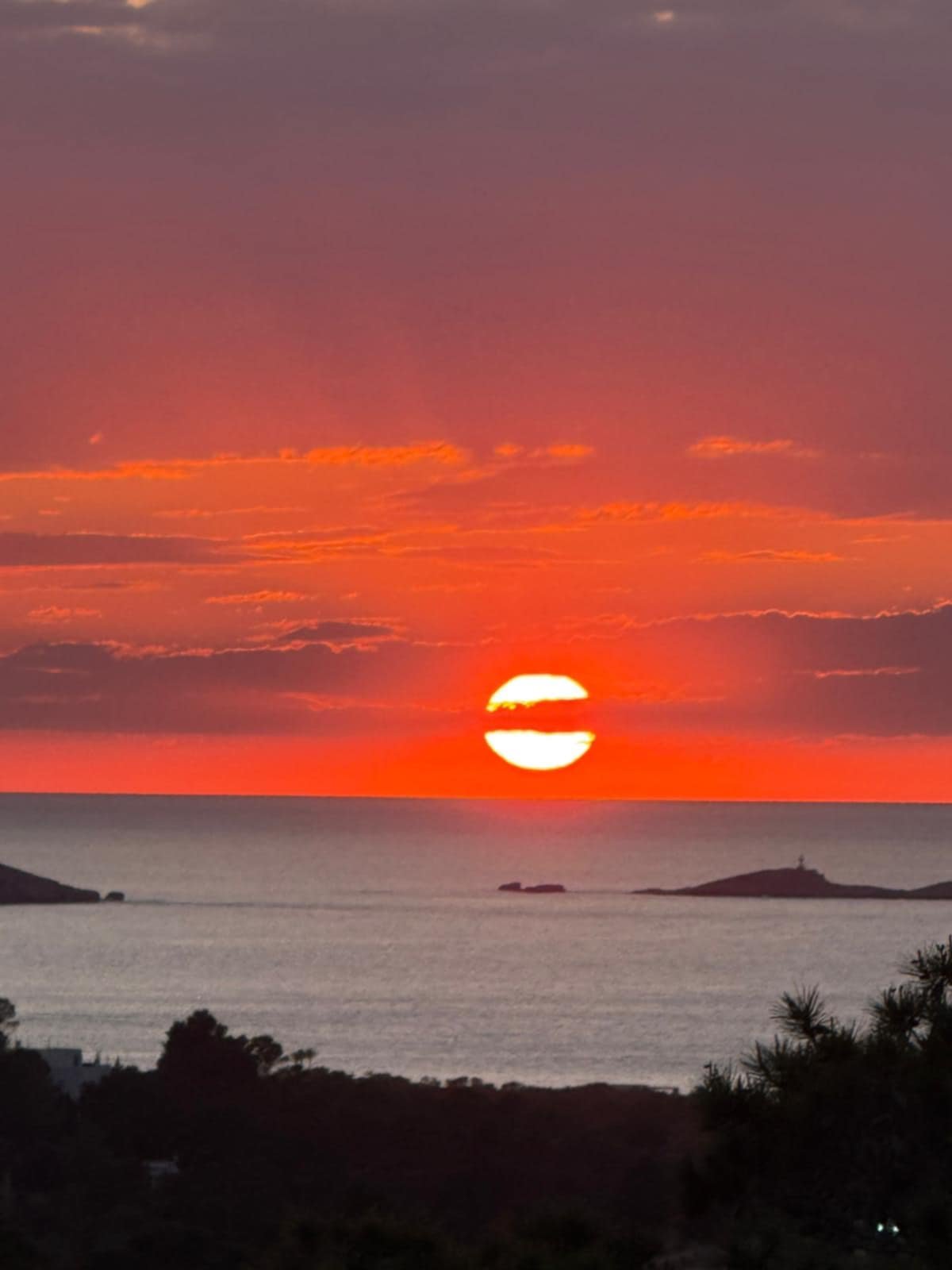Sunset over the ocean with a partially obscured sun and a colorful sky, including clouds and a silhouette of islands and trees in the foreground.