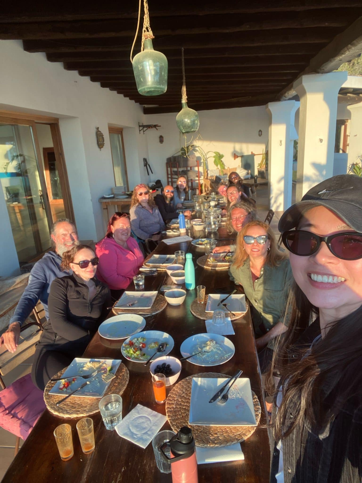 A group of people sitting around a dining table outdoors, smiling and enjoying a meal together during sunset.