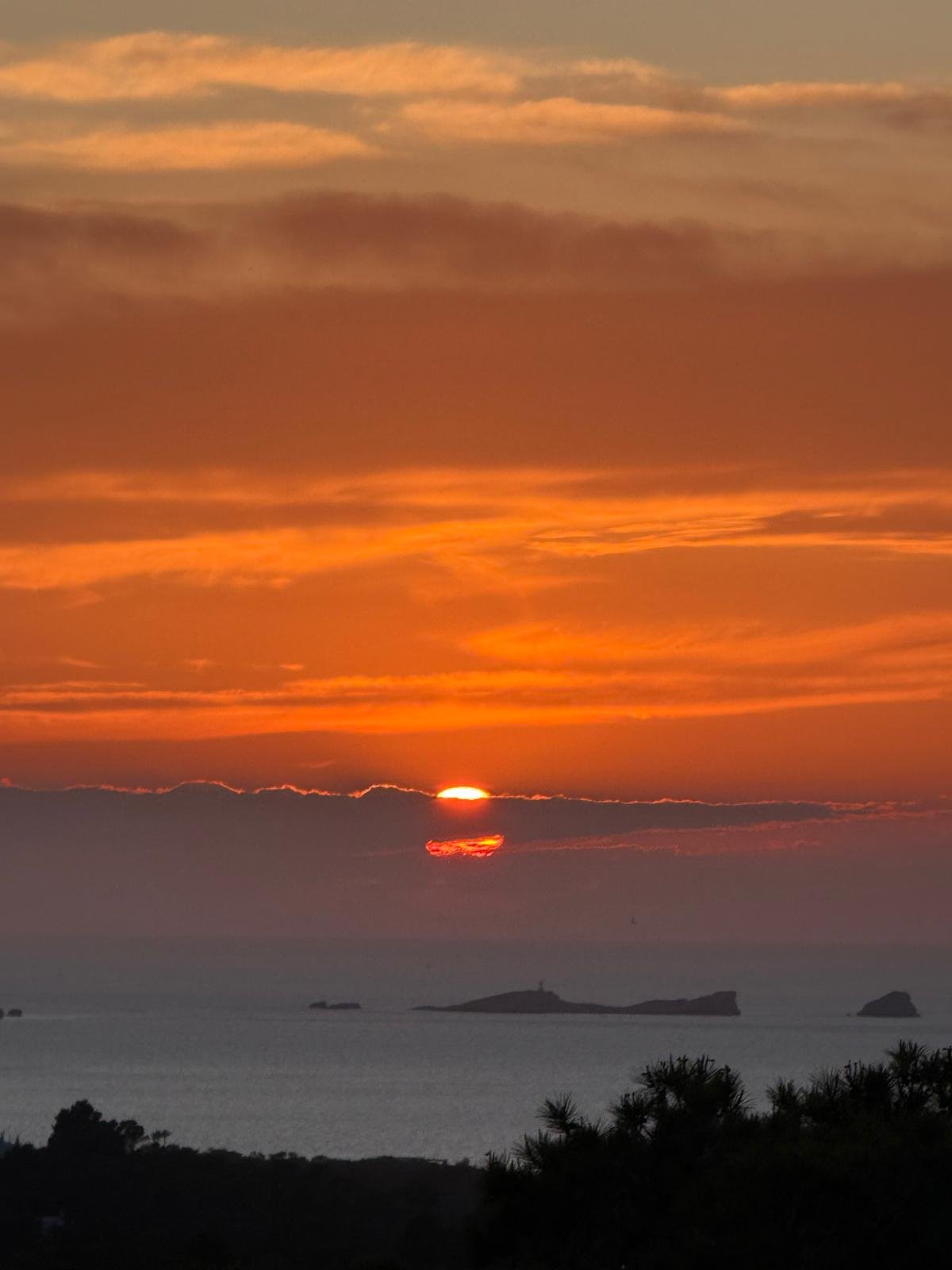 Sunset over the ocean with partially obscured sun and distant islands, silhouettes of trees in the foreground.