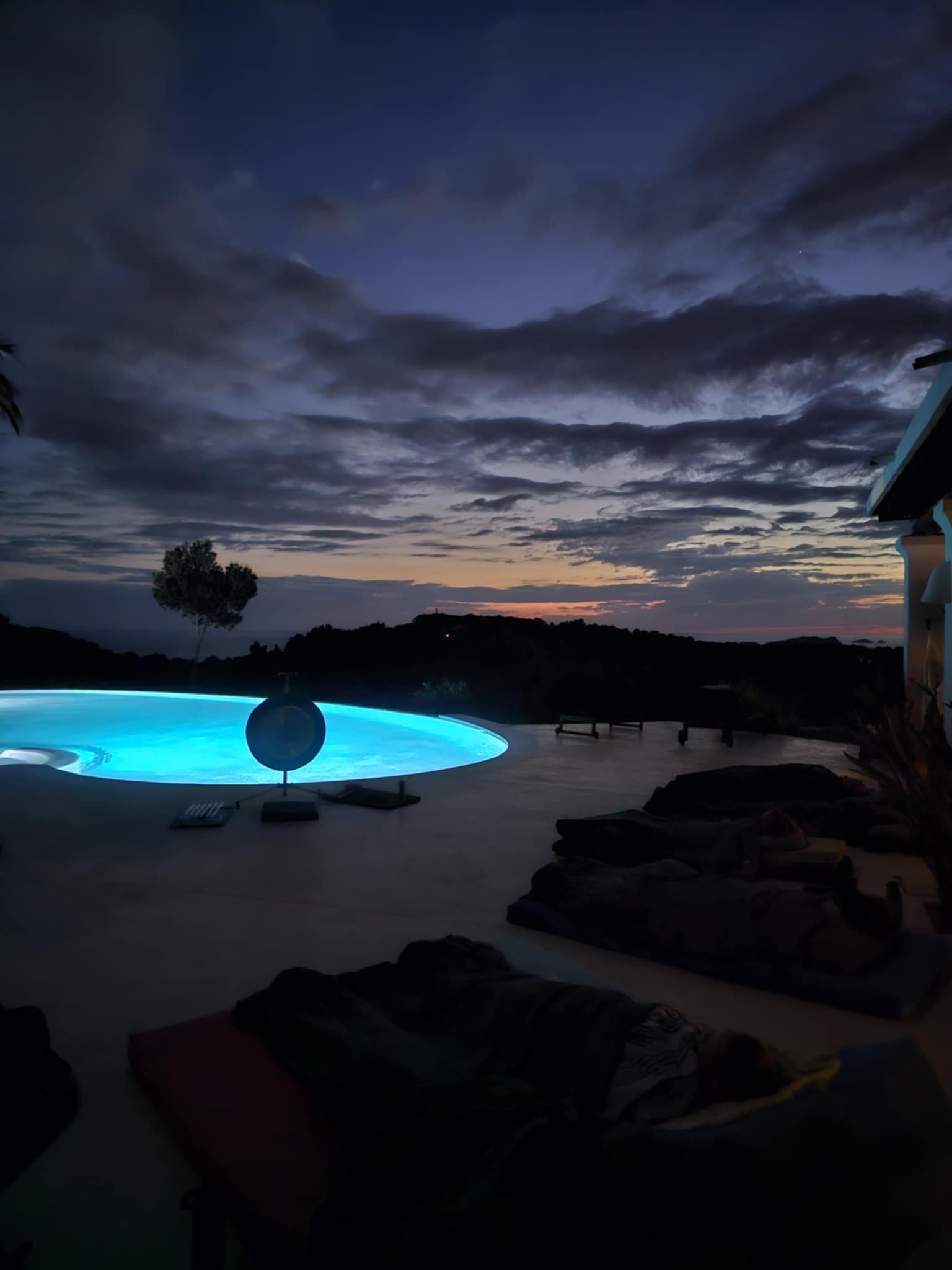 People lying on lounge chairs near a lit-up swimming pool at dusk with dark cloudy sky and distant hills in the background.