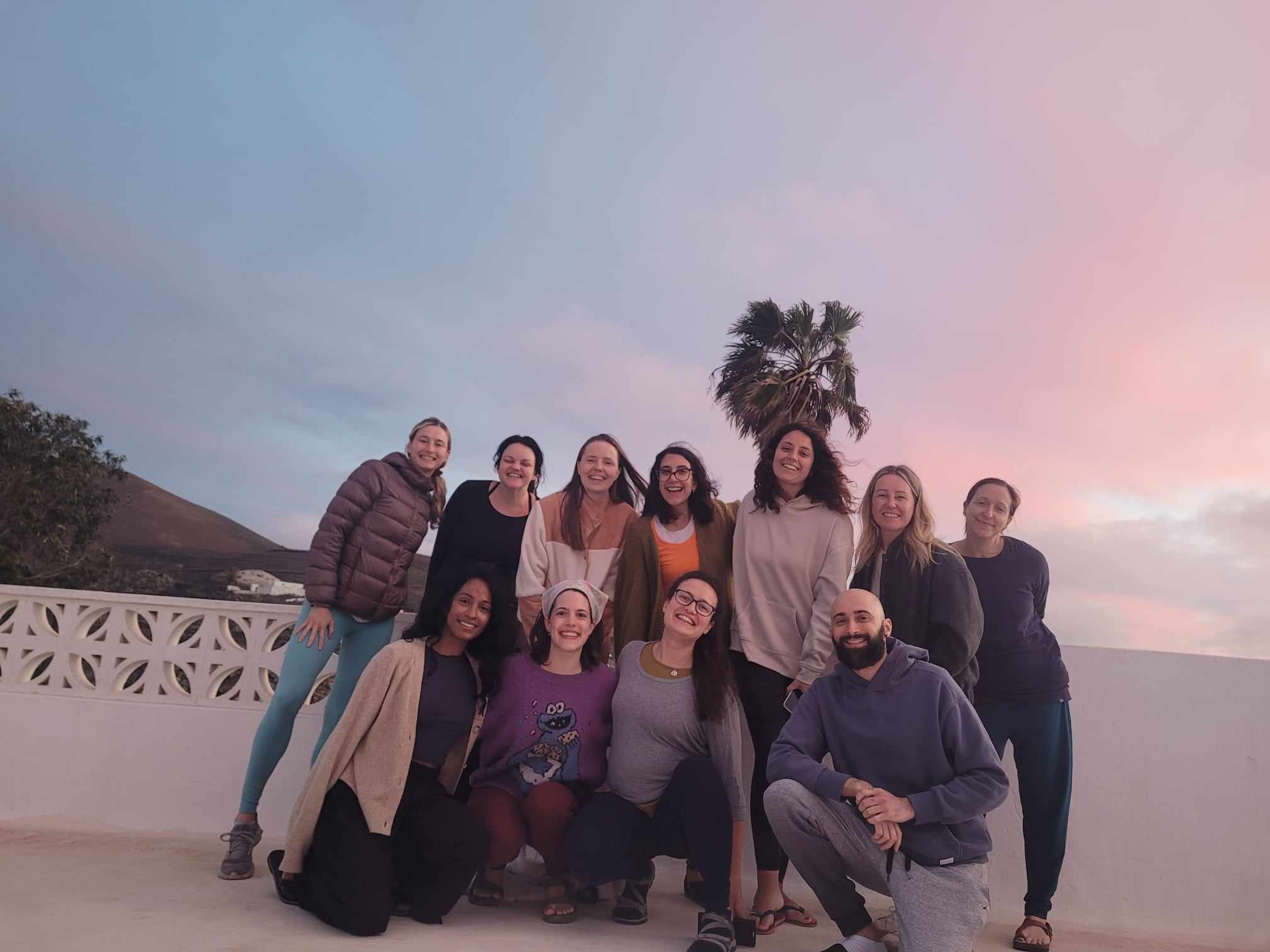 Group of eleven people smiling outdoors during sunset, standing and kneeling on a white terrace with a palm tree and a hilly landscape in the background.