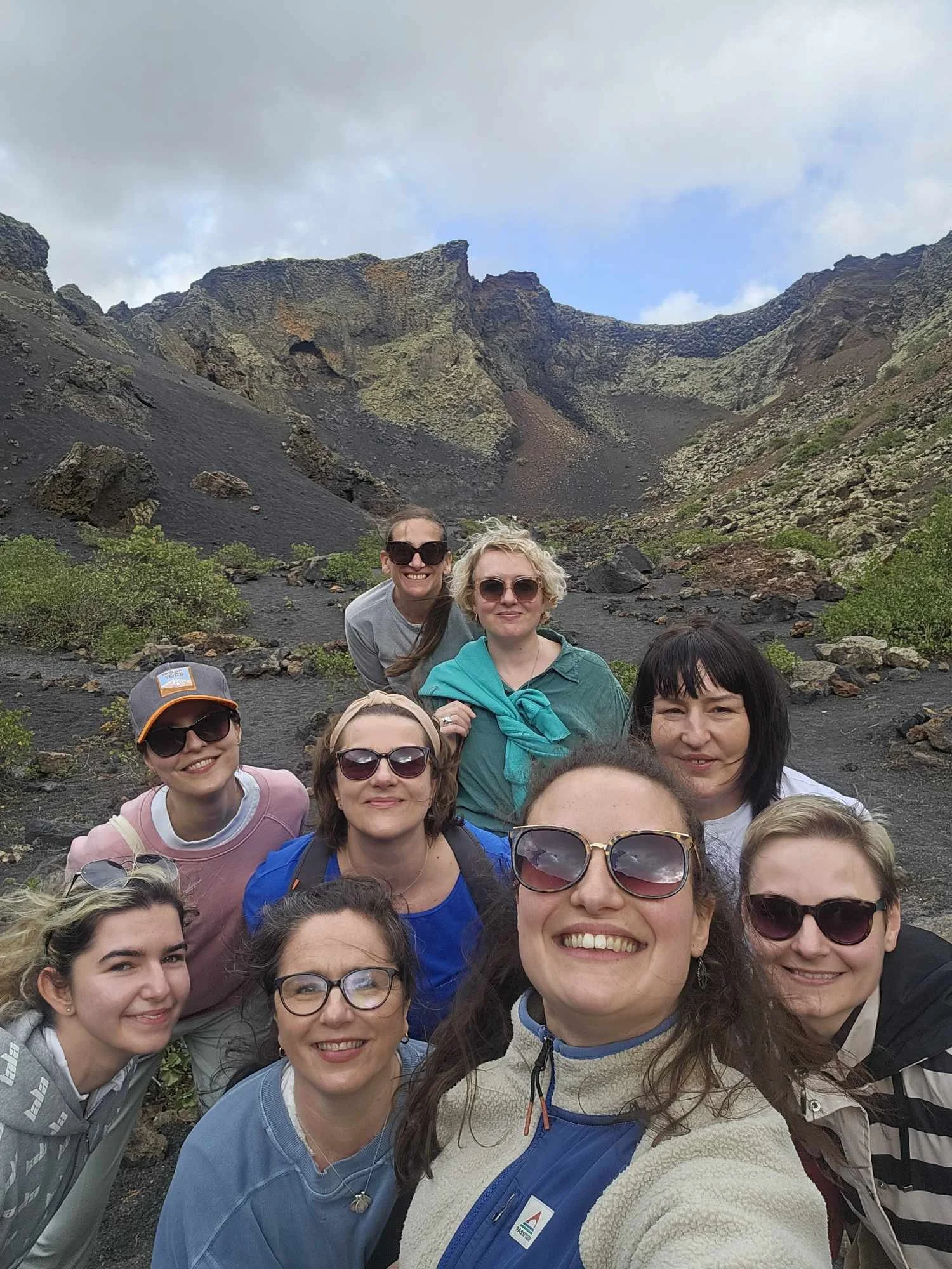Group of ten women smiling and taking a selfie in a volcanic landscape with dark rocks and steep cliffs.