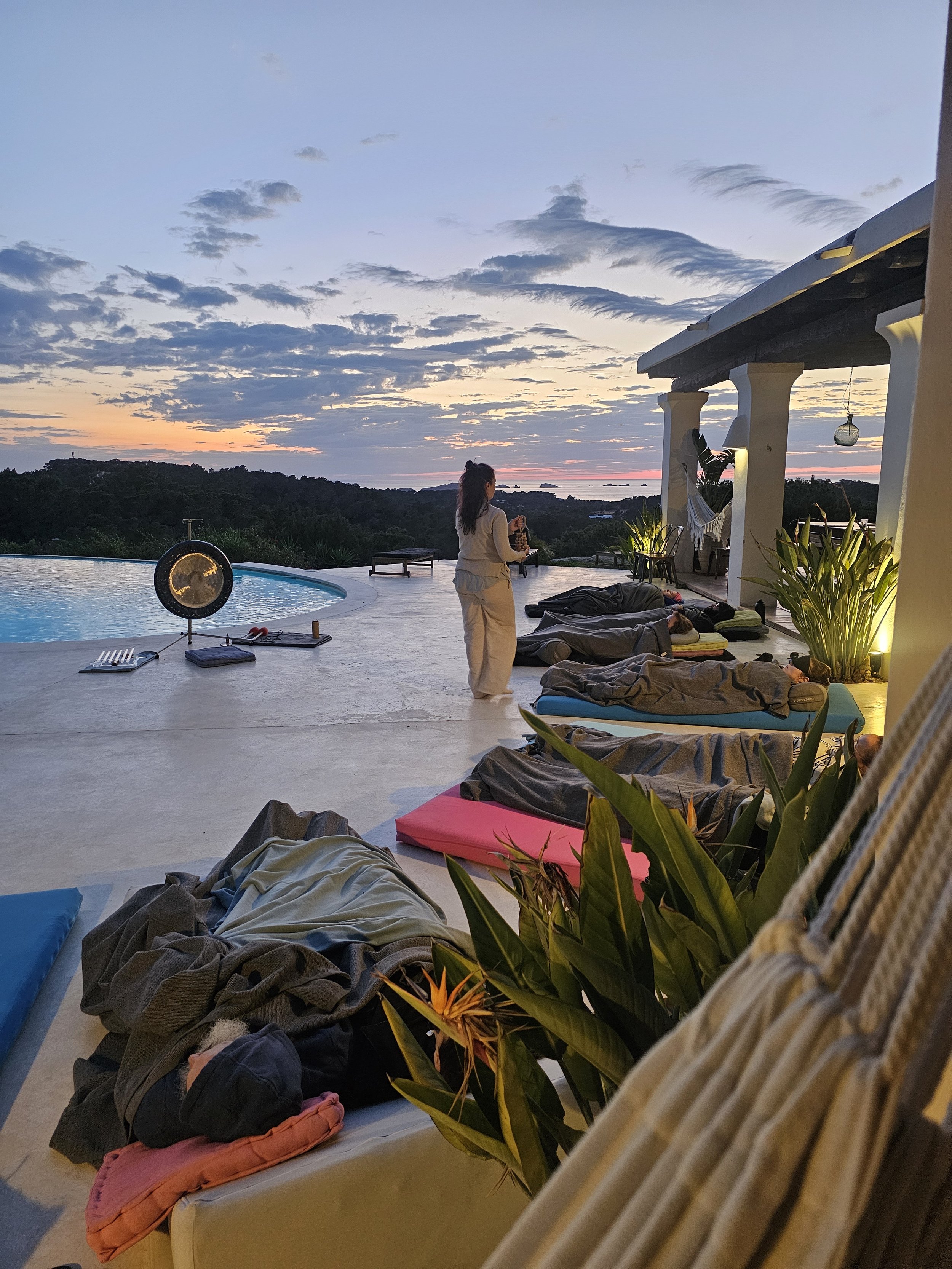People relaxing on mats and cushions by an infinity pool during sunset with a woman standing and holding a beverage, overlooking a scenic view of greenery and the ocean.