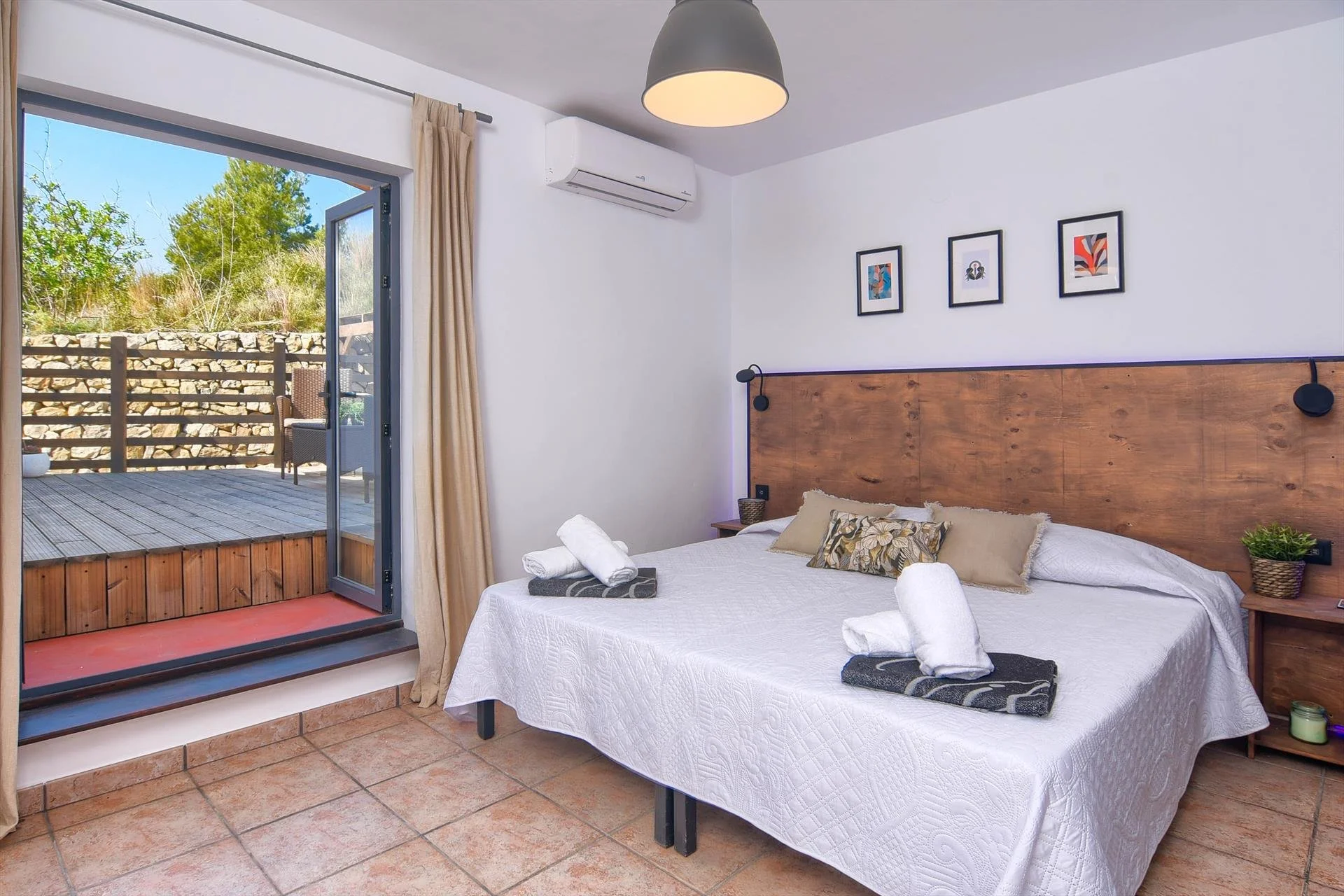 Bedroom with a white bedspread, wooden headboard, artwork above, and a sliding glass door open to a wooden deck with outdoor seating and a stone wall.