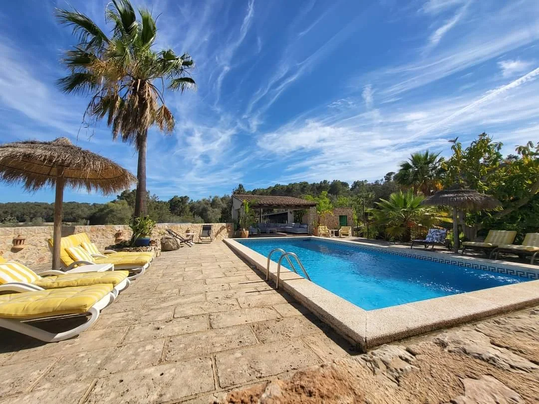 Sunny outdoor swimming pool with blue water, surrounded by yellow and white lounge chairs, straw umbrellas, palm trees, and lush greenery under a partly cloudy blue sky.