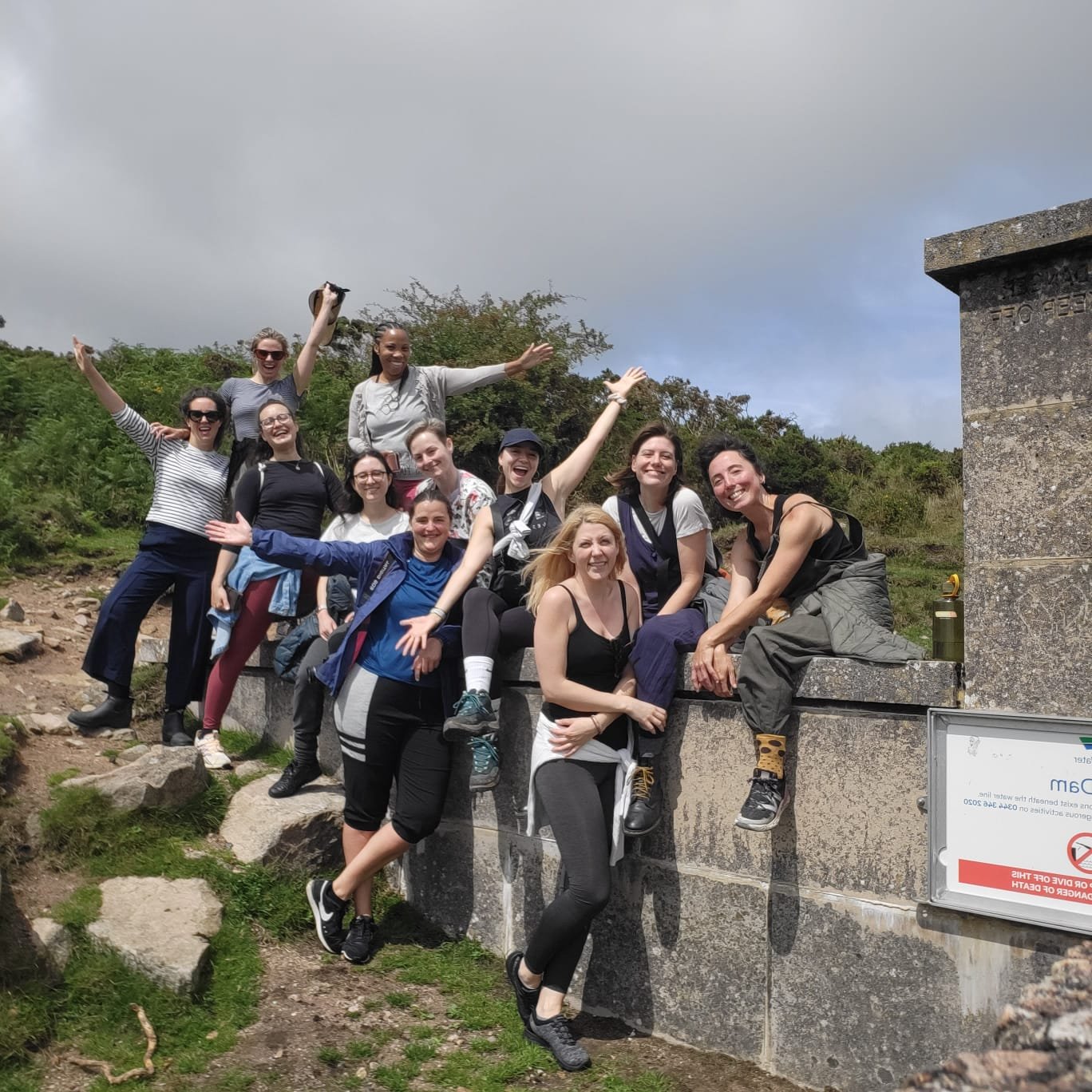 Group of friends outdoors on a sunny day, sitting and standing on a stone wall, smiling and celebrating.
