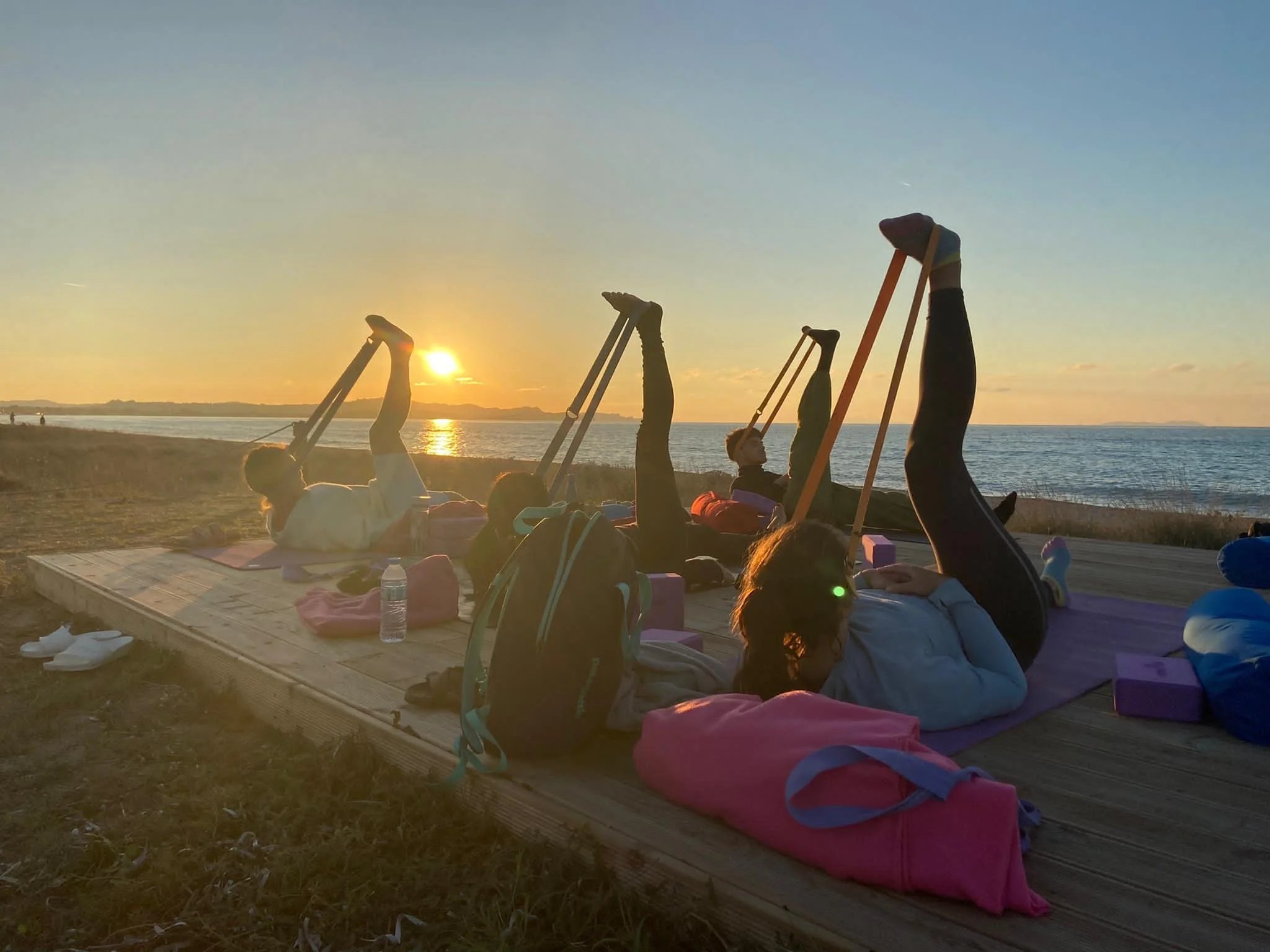Group of people practicing yoga on a wooden platform by the beach at sunset, with water and the horizon in the background.